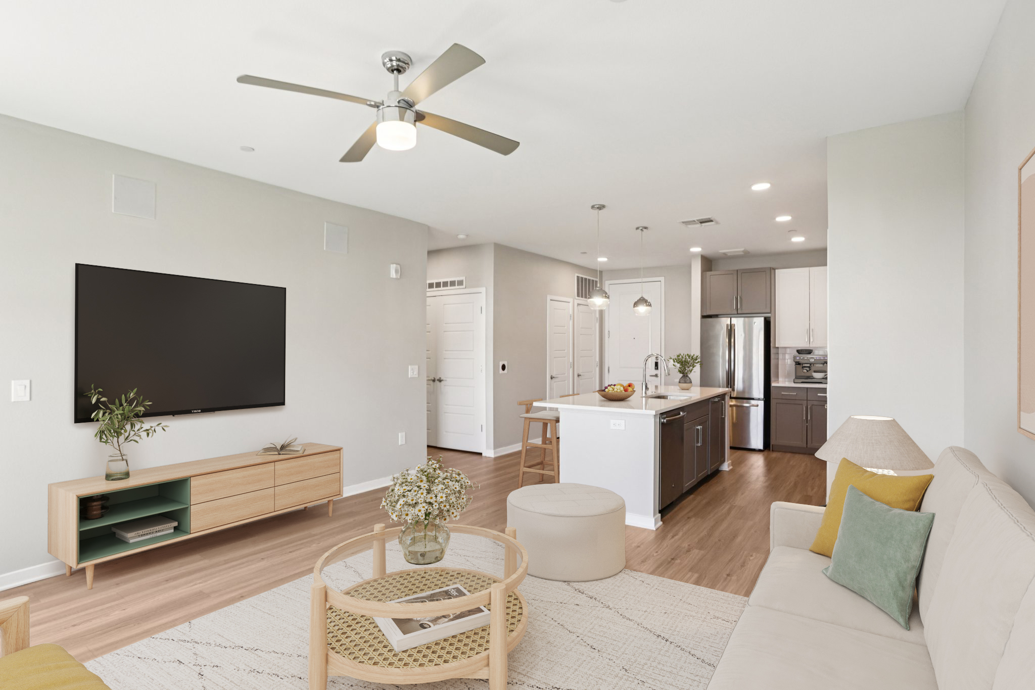 A modern living room featuring a beige sofa with decorative pillows, a round coffee table, and a large flat-screen TV mounted on the wall. The open-concept design leads to a kitchen with sleek cabinetry, stainless steel appliances, and a central island. Bright, natural light fills the space through large windows.