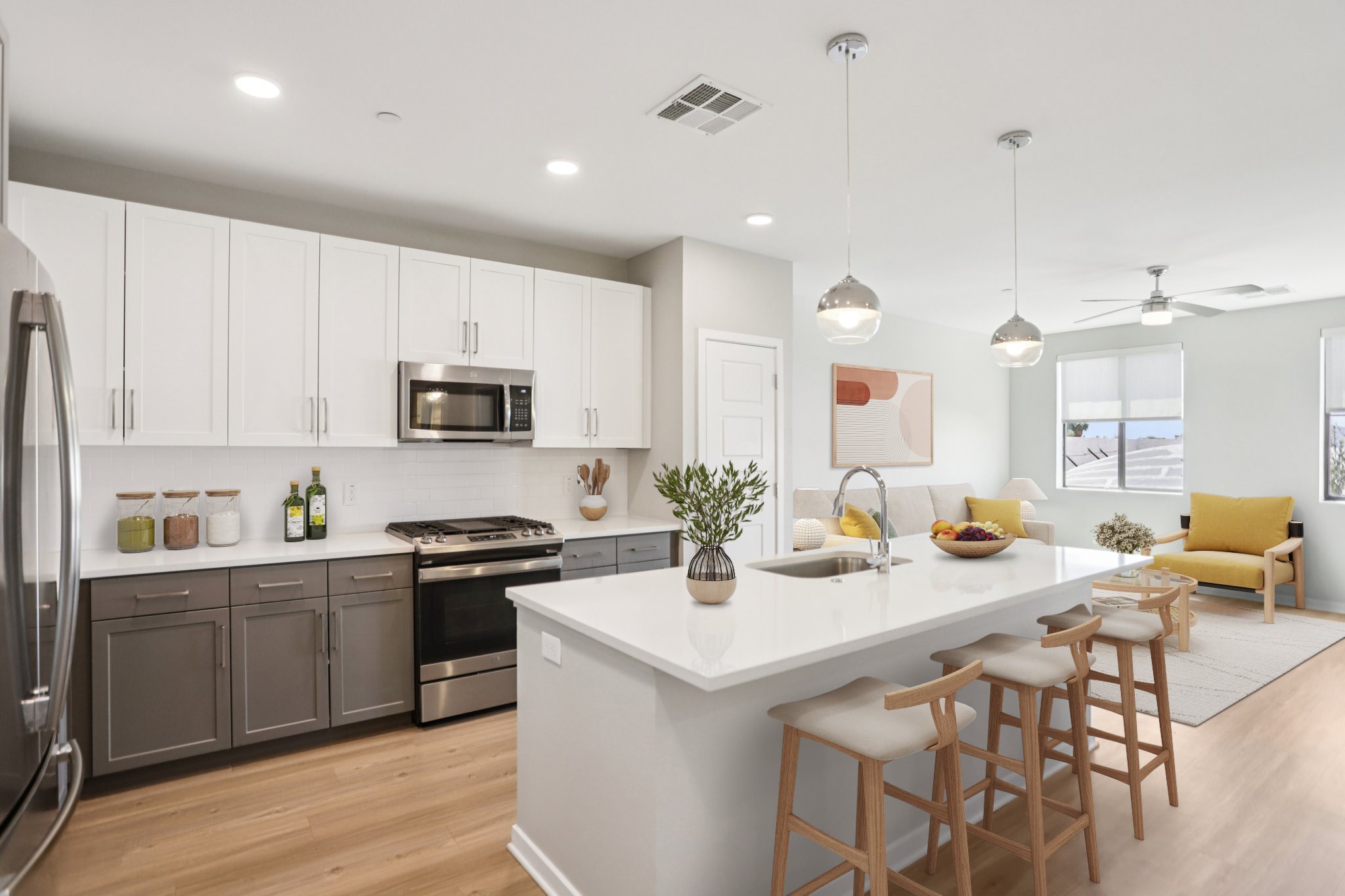 A modern kitchen with white and gray cabinetry, stainless steel appliances, and a large island with bar stools. The space features natural lighting, a living area with a yellow chair, and decorative elements like a bowl of fruit and a potted plant.