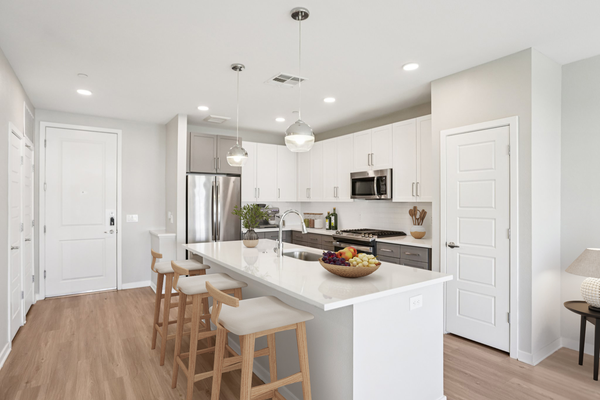 Modern kitchen featuring a large island with three stools, stainless steel appliances, white cabinetry, and a spacious layout. Natural light fills the room, highlighting a bowl of fruit on the counter and decorative plants. Light wood flooring adds warmth to the contemporary design.