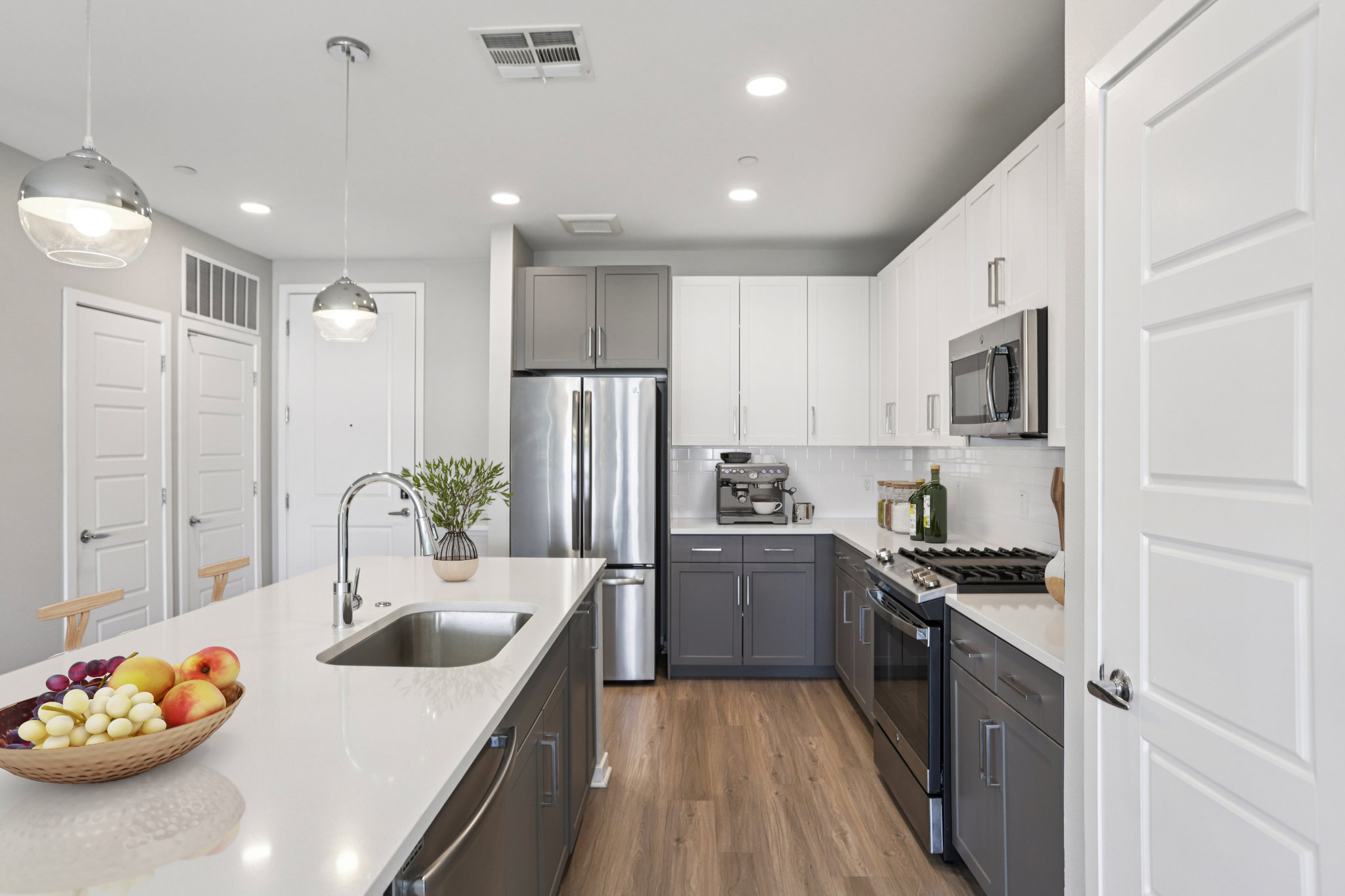 Modern kitchen featuring sleek gray and white cabinetry, stainless steel appliances, and a large island with a sink. A bowl of colorful fruit sits on the counter, and pendant lights illuminate the space. The walls are neutral-colored, creating a bright and inviting atmosphere.