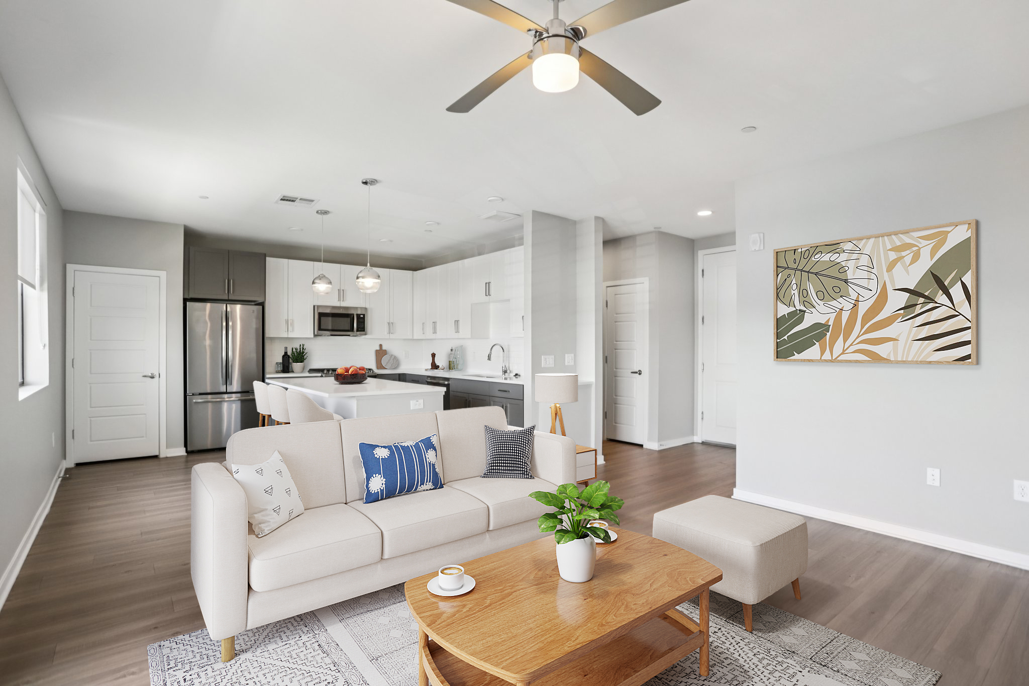 A modern living room with a light-colored sofa adorned with decorative pillows, a wooden coffee table, and a potted plant. In the background, there's a bright kitchen featuring white cabinets, stainless steel appliances, and an island. Soft lighting and neutral walls create a cozy atmosphere.