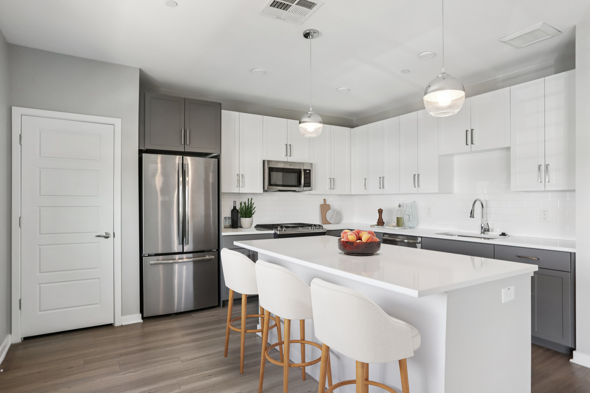 Modern kitchen featuring sleek gray and white cabinetry, stainless steel appliances, and a large island with seating. The countertop is adorned with a bowl of apples, and the space is well-lit with pendant lighting, showcasing a clean, contemporary design.