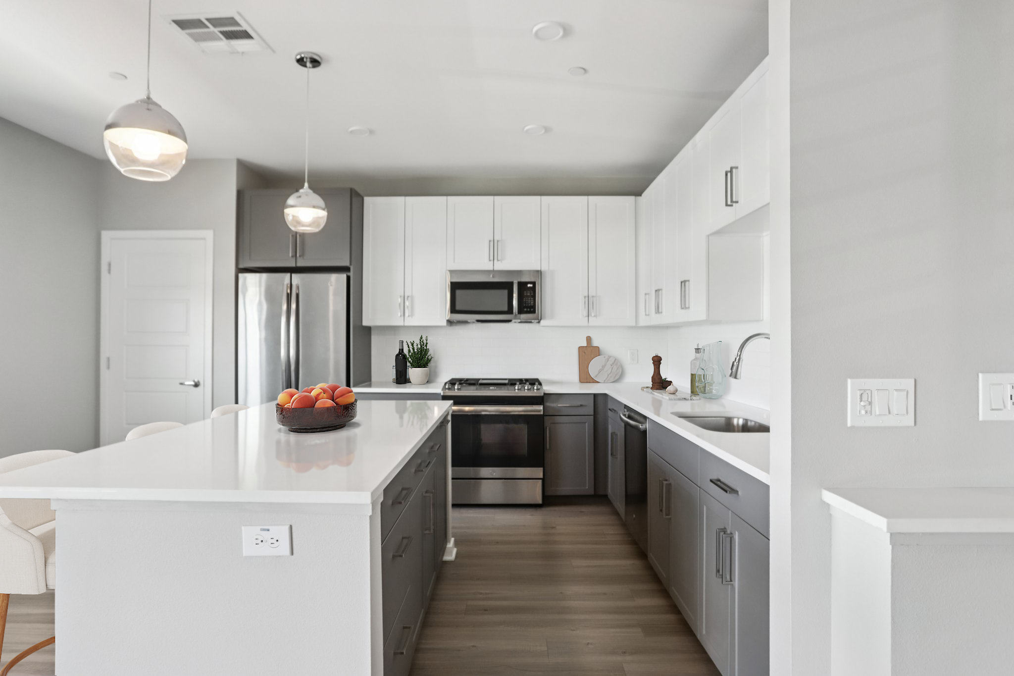 Modern kitchen featuring a central island with a bowl of oranges, sleek white upper cabinets, and gray lower cabinets. Stainless steel appliances include a refrigerator, microwave, and stove. The space is well-lit with pendant lighting and has a clean, minimalist aesthetic.