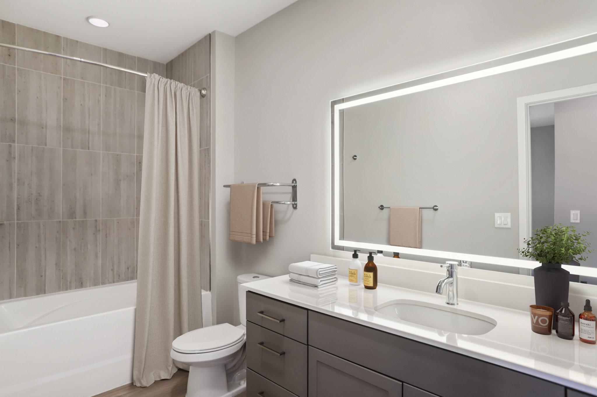 A modern bathroom featuring a tub with a beige shower curtain, a large mirror with backlighting, and a sleek vanity. The vanity has dark wood cabinetry, a white countertop with toiletries, and a small potted plant. Neutral-colored walls and flooring create a contemporary feel.
