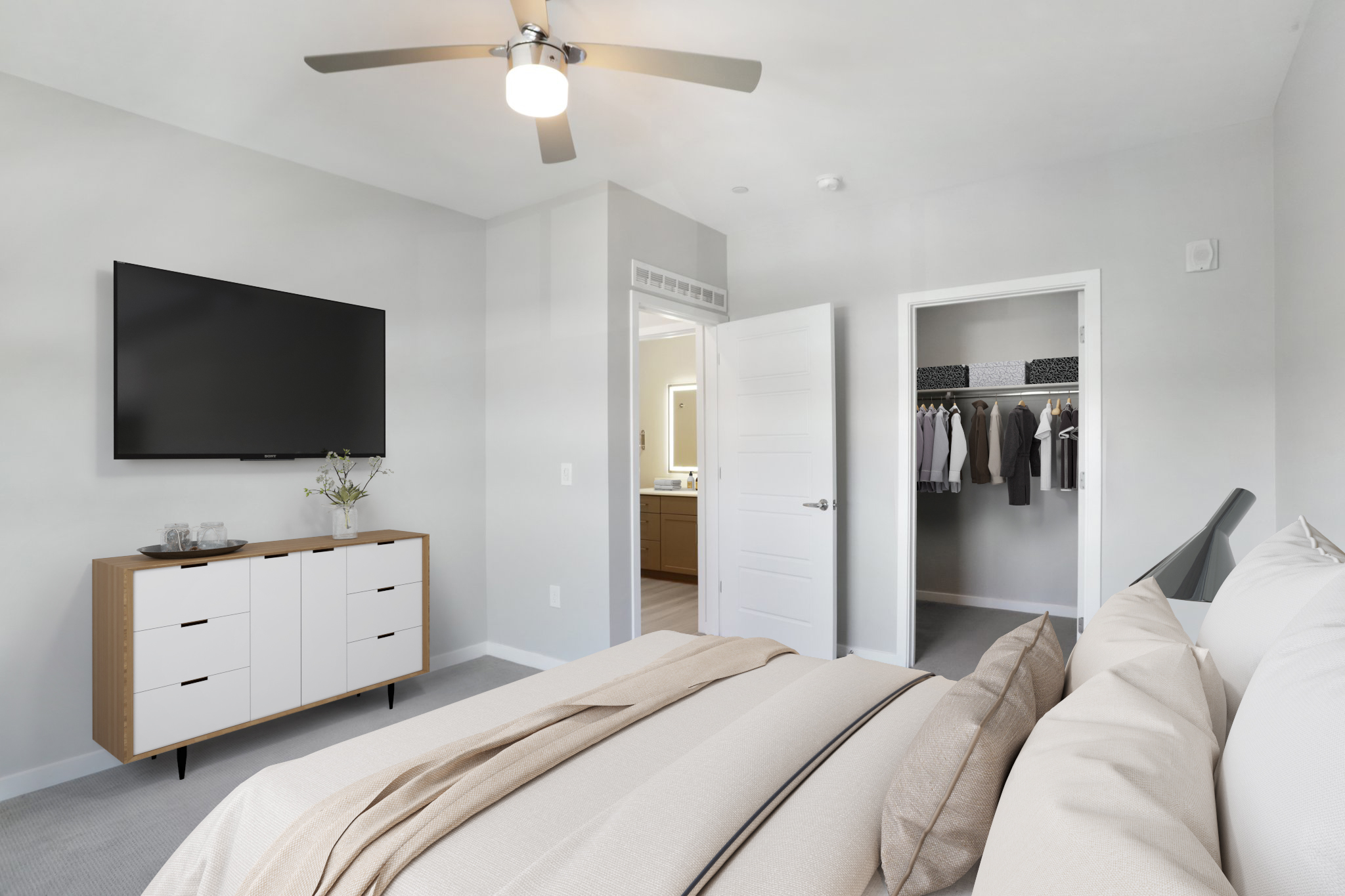 A modern bedroom featuring a cozy bed with light-colored bedding, a ceiling fan, a wall-mounted TV, and a light-colored dresser. A closet door is open to reveal organized clothing. The walls are painted in soft gray tones, and there is natural light coming from an adjacent room.