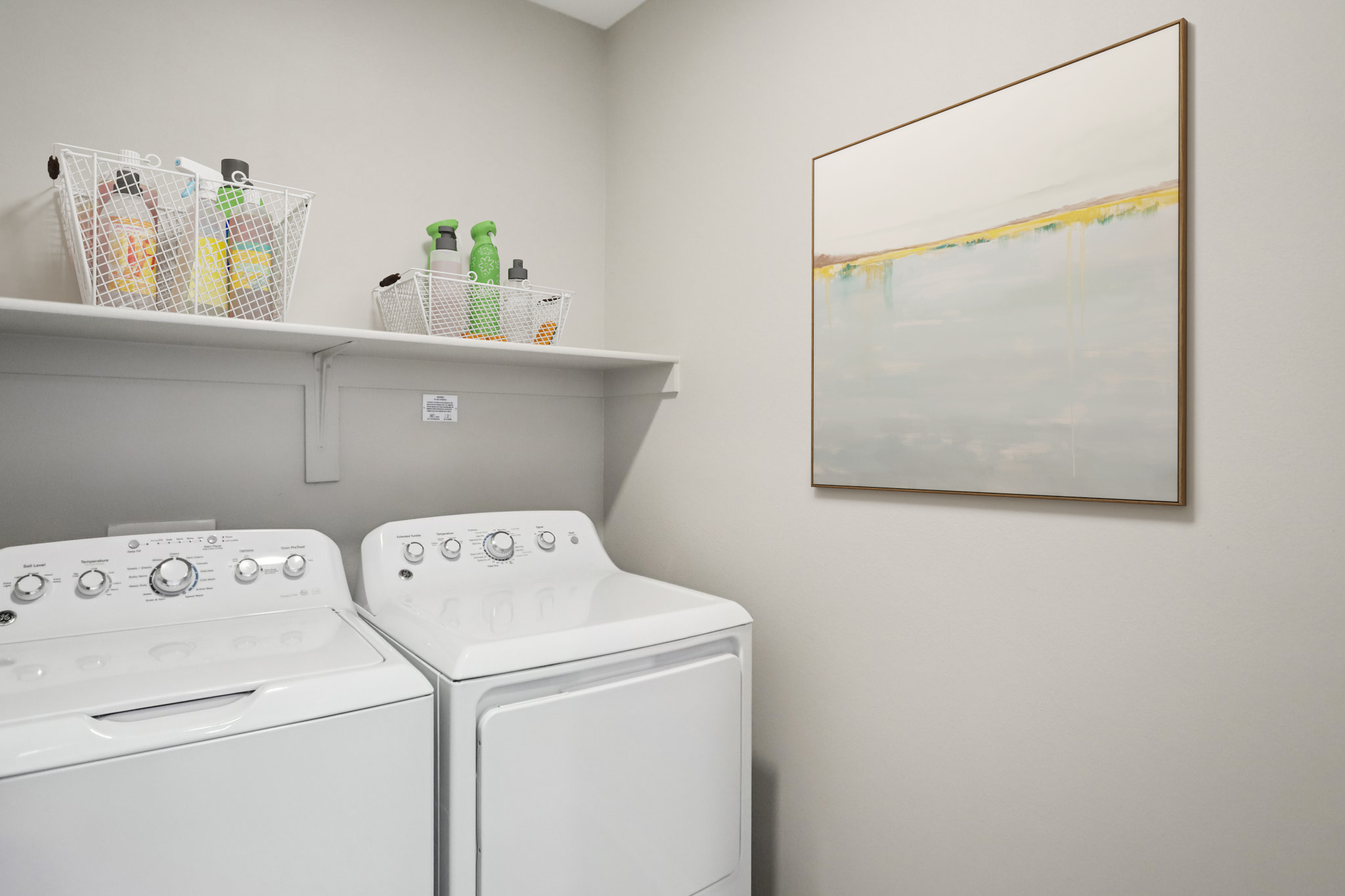 A laundry room featuring a washer and dryer side by side. Above them, a shelf holds several baskets with cleaning supplies. A large abstract painting hangs on the wall, adding a touch of style to the functional space. The walls are painted in a soft, neutral color.