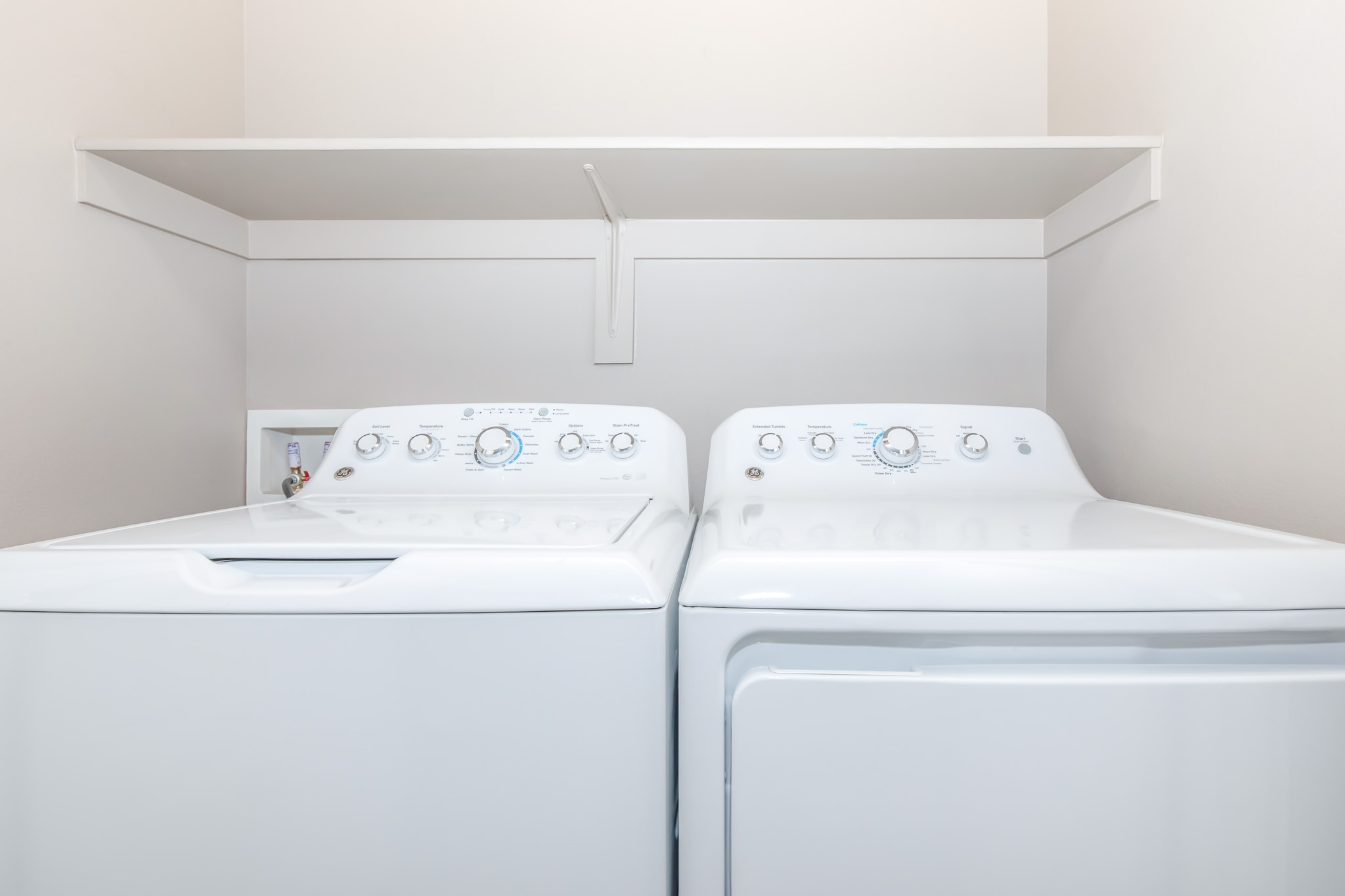 A clean laundry room featuring a stacked set of white washing machine and dryer. The appliances are positioned side by side against a light-colored wall, with a shelf above them for storage. The controls on the machines are visible, highlighting their modern design.