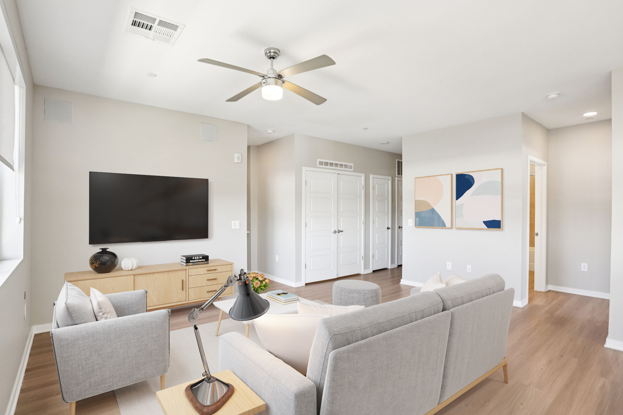 A modern living room featuring a light gray sofa and armchair, a wooden coffee table, and a TV mounted on the wall. The space is well-lit with a ceiling fan and has two abstract wall art pieces. Light beige walls and wooden flooring create a cozy atmosphere.