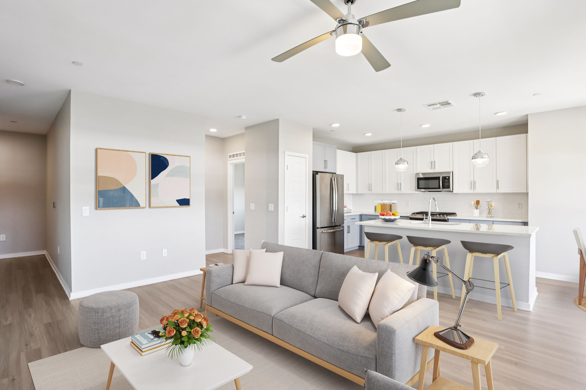 A modern, open-concept living space featuring a light gray sofa with white pillows, a coffee table with a vase of flowers, and art on the wall. The kitchen has white cabinets, stainless steel appliances, and a bar with stools. Natural light fills the room, highlighting the clean, contemporary design.