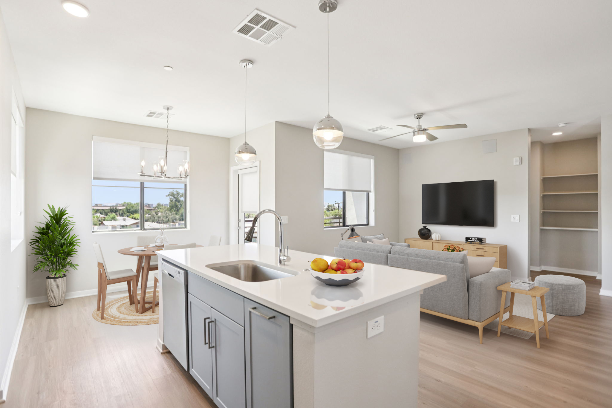 Modern open-concept living space featuring a kitchen island with fruit, a dining area with a round table, and a cozy living room with a television. Large windows provide natural light, and contemporary furnishings create an inviting atmosphere. A green plant adds a touch of nature to the decor.