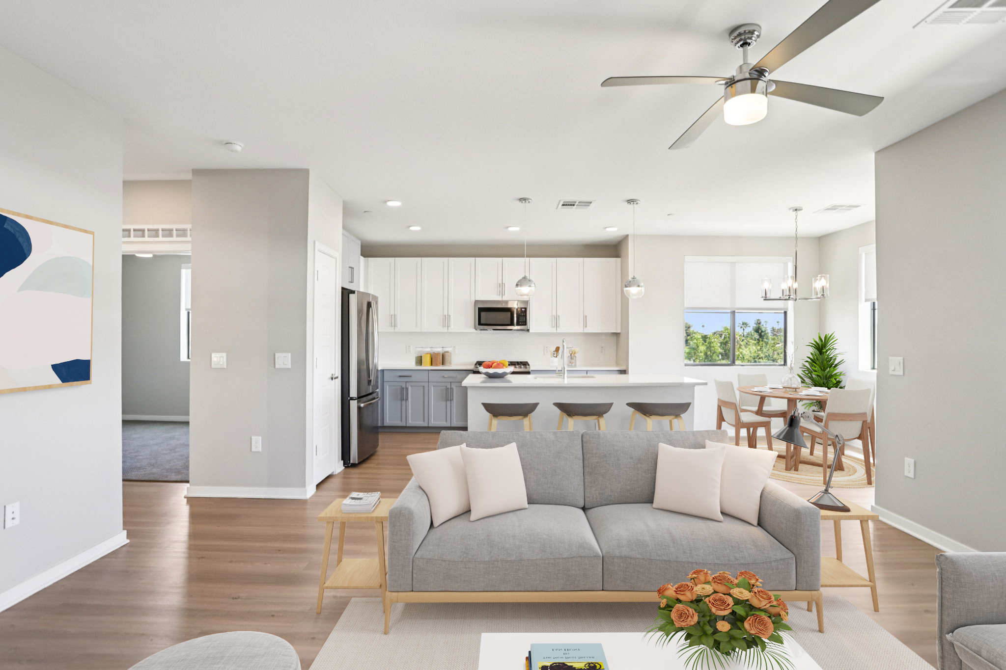A modern, open-concept living area featuring a gray sofa with decorative pillows, a coffee table with flowers, and a dining space with wooden chairs. The kitchen has white cabinetry and stainless steel appliances, with bright natural light coming through the windows and a ceiling fan above.