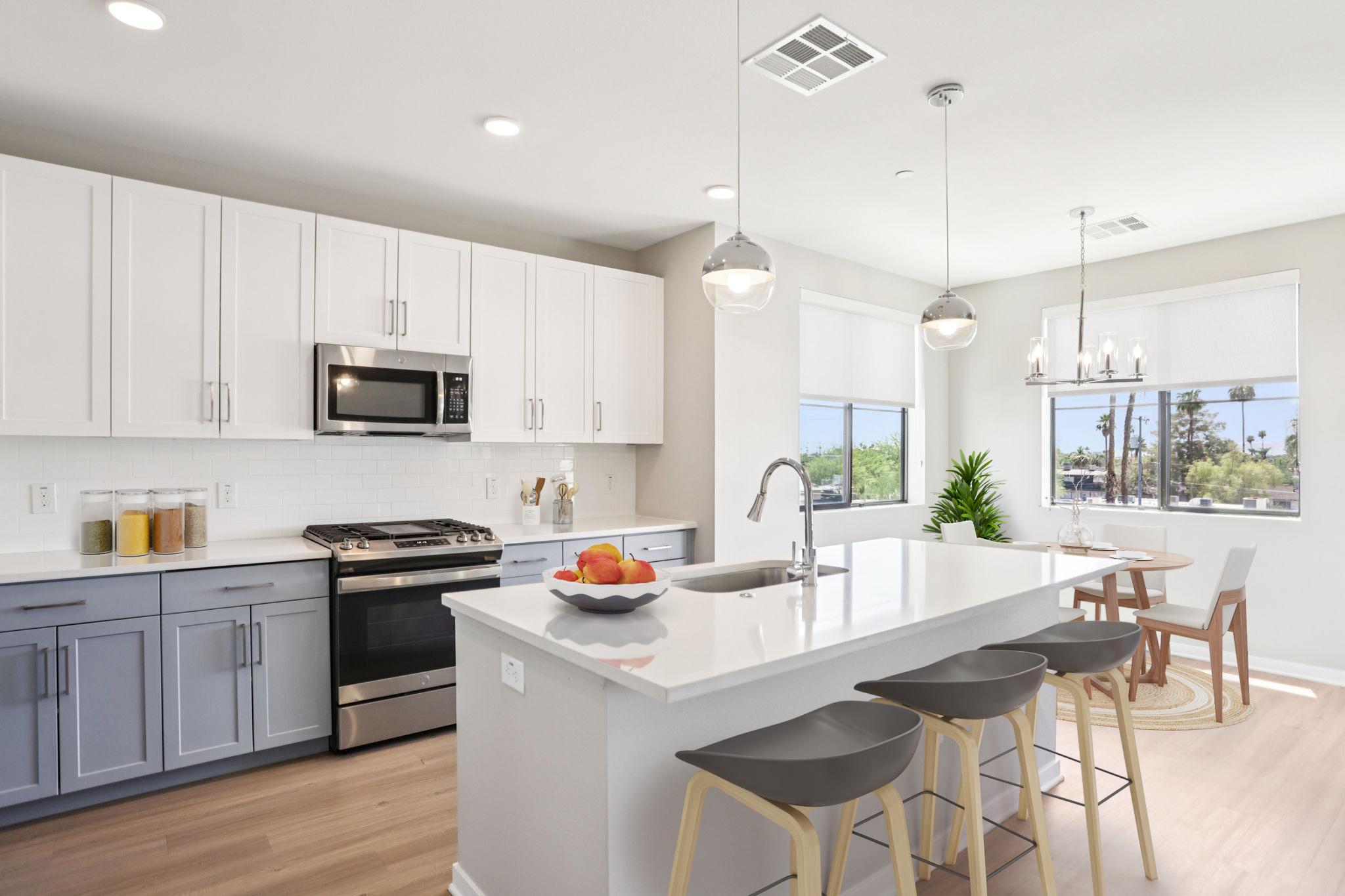 A modern kitchen with white cabinets and blue lower cabinets. The kitchen features a stainless steel stove and microwave, a large island with a sink, and three bar stools. There is a bowl of fruit on the island, and a bright dining area with a table and chairs beside large windows that provide natural light.