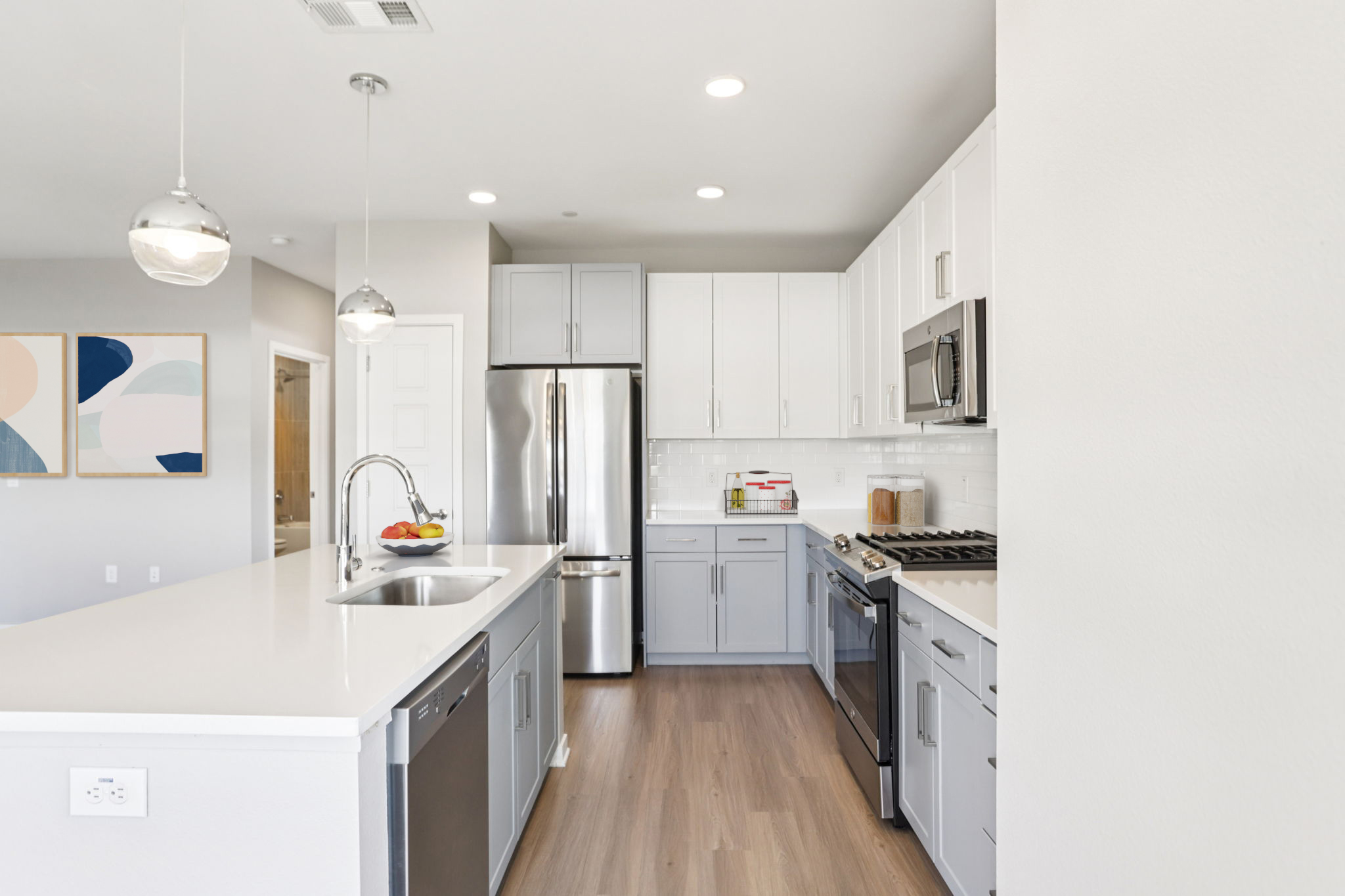 Contemporary kitchen featuring a central island with a sink, white countertops, stainless steel appliances, and a mix of light gray and white cabinetry. Pendant lighting hangs above the island, with a bowl of fruit for decor. The space has a modern, open layout with natural lighting.