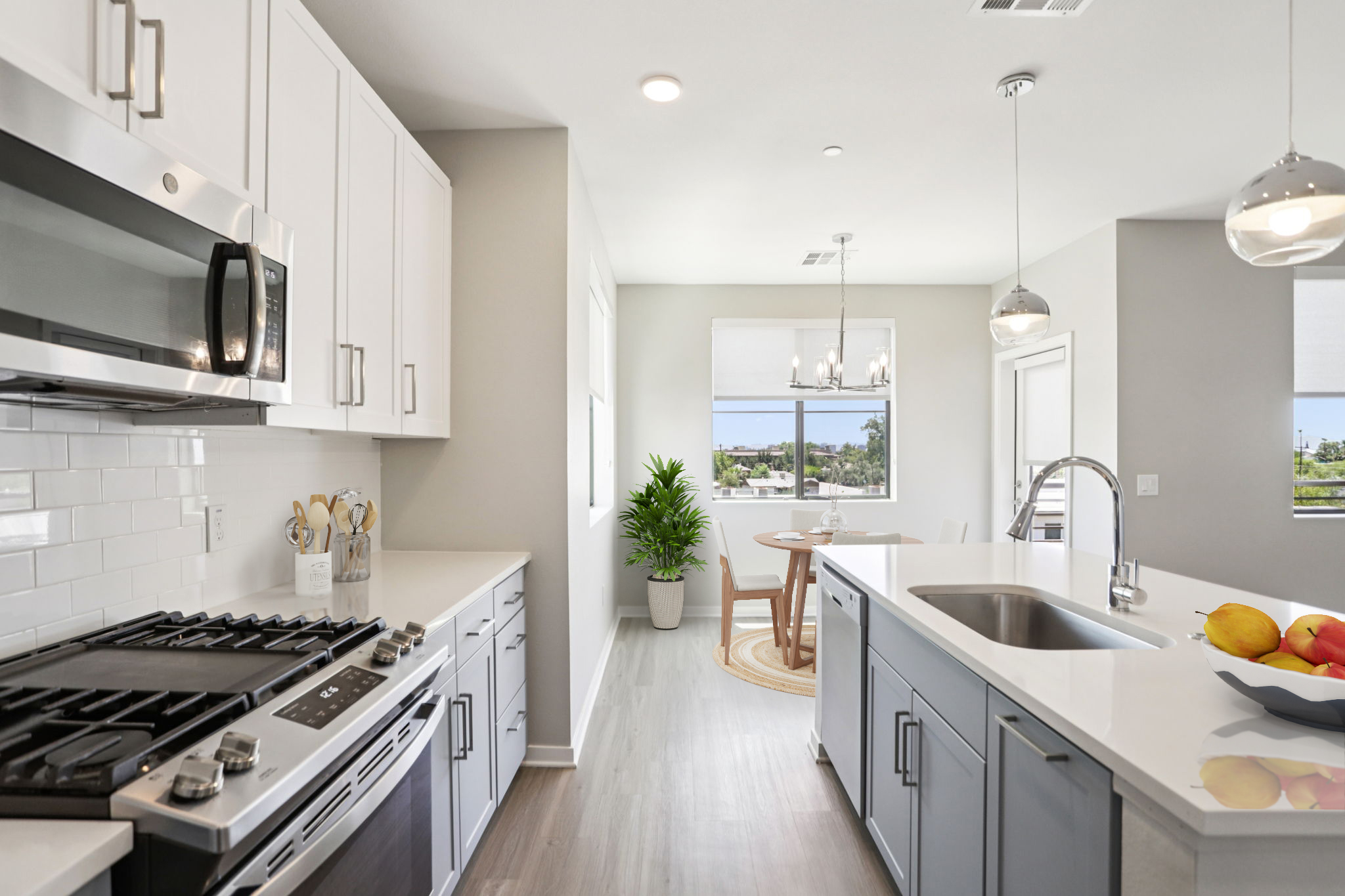 Modern kitchen with white cabinets and gray lower cabinets, featuring stainless steel appliances, a gas stove, and a large sink. Natural light streams in from a nearby window, illuminating a small dining area with a round table and chairs, complemented by a decorative plant and a bowl of fruit on the countertop.