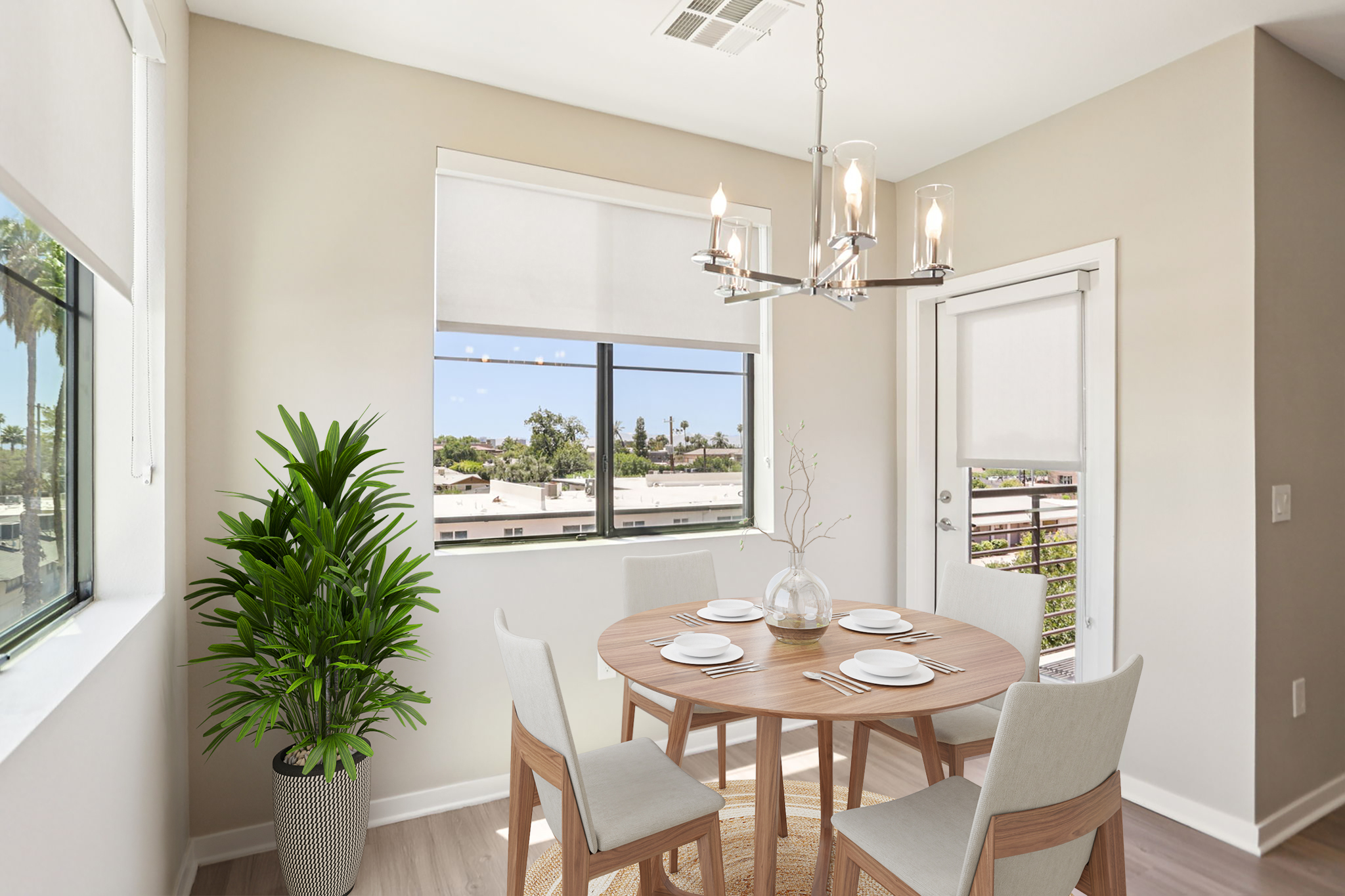 A modern dining area featuring a round wooden table set with four white plates. Light-colored chairs surround the table, and a decorative plant is positioned in the corner. Large windows offer a view of greenery outside, and a stylish chandelier hangs overhead, enhancing the bright, airy atmosphere.