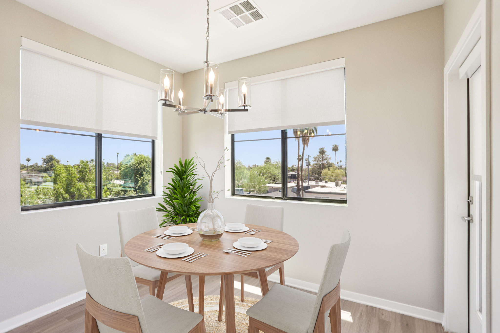 A modern dining area featuring a round wooden table set for four with plates and utensils. The space is illuminated by a stylish chandelier and has large windows with views of greenery outside. Light-colored walls and flooring create a bright, inviting atmosphere.
