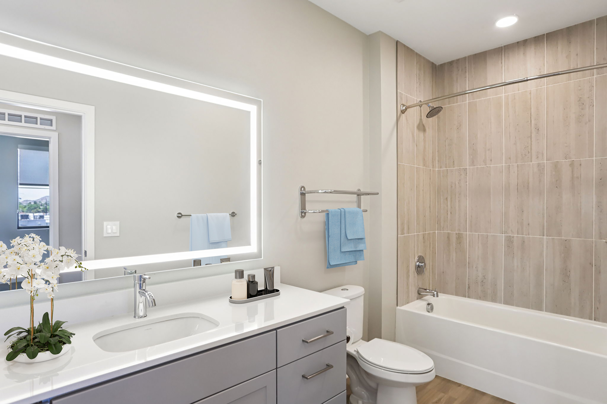 A modern bathroom featuring a light vanity with a sink, a large mirror with backlighting, a bathtub with a shower, and a towel rack. Soft blue towels add a pop of color, and a decorative plant sits on the counter for a touch of nature. The walls are styled with light wooden tiles.