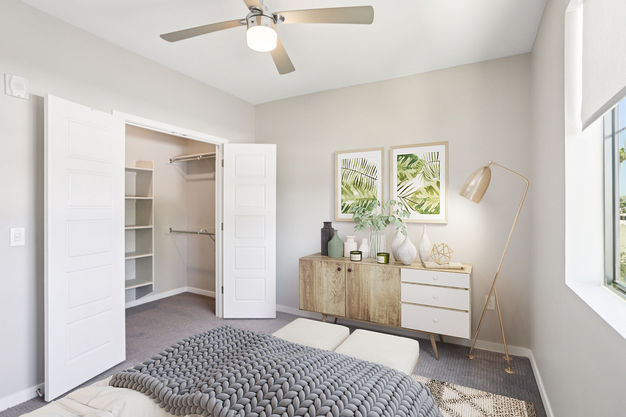 A bright and modern bedroom featuring a cozy bed with a textured blanket, a stylish dresser with decorative elements, an open closet with shelves, and framed botanical art on the wall. A ceiling fan and large window provide light and airiness to the space.