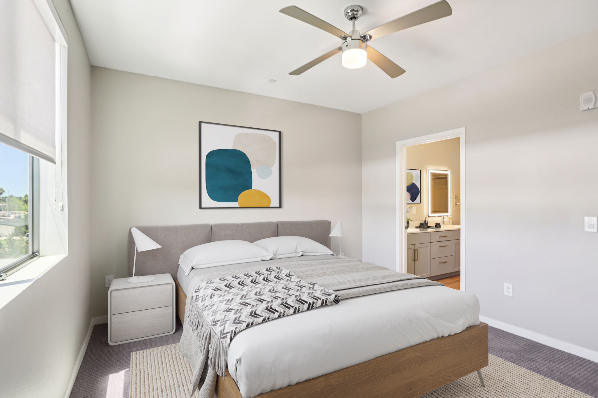 A modern bedroom featuring a double bed with a light gray headboard and a patterned throw blanket. There is a bedside table with a minimalist lamp, a large window allowing natural light, and a framed abstract artwork on the wall. A doorway leads to a bathroom in the background.
