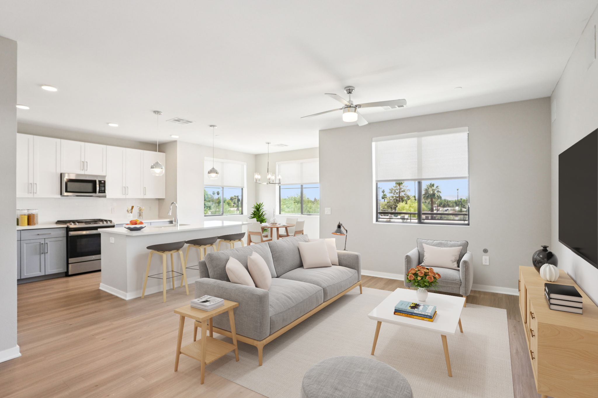 A modern, open-concept living room and kitchen area featuring light-colored walls, wooden flooring, and large windows. The living room includes a gray sofa, a light coffee table, and a decorative plant. The kitchen has white cabinets, stainless steel appliances, and a breakfast bar with stools. Natural light fills the space.