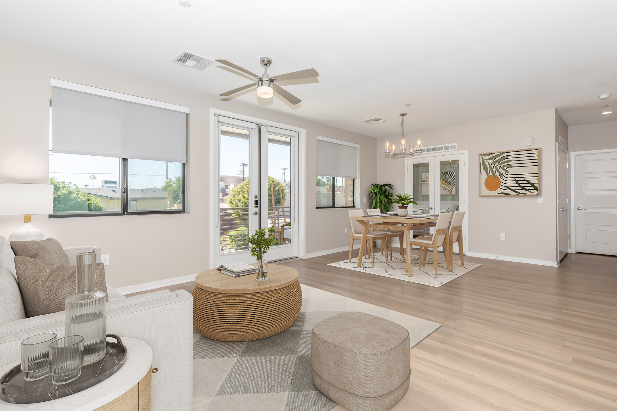 A modern and spacious living room featuring neutral tones. A comfortable sofa and a round coffee table are in the foreground. There are large windows with light filtering shades, a dining area with a wooden table and chairs, and decorative plants. The space is well-lit and inviting, showcasing contemporary design elements.
