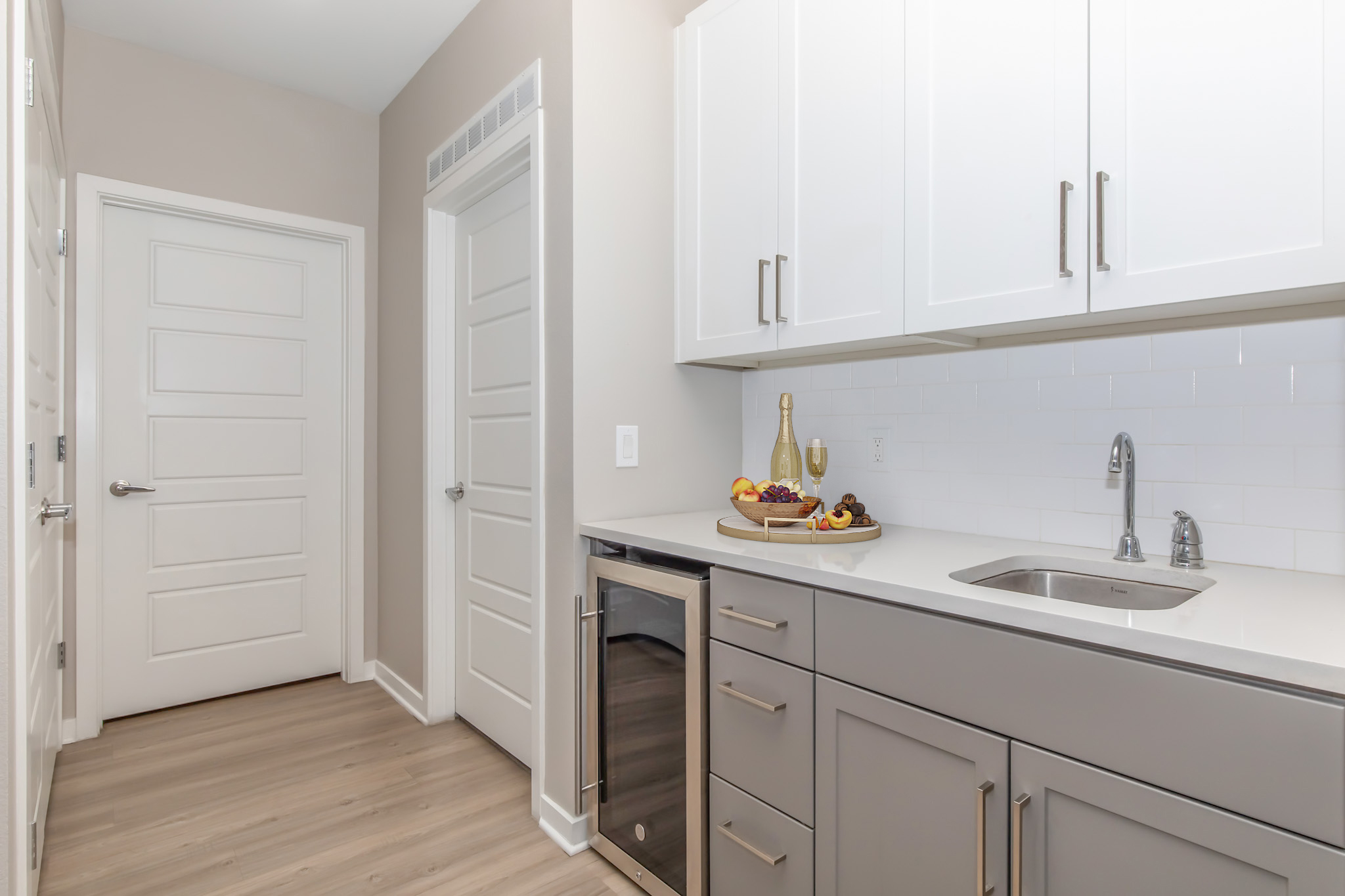 A modern kitchen space featuring gray cabinets and white countertops, with a sink and a small wine fridge. A decorative tray with fruit is placed on the counter. There are closed doors on the left and right sides, and the walls are painted a light neutral color. The flooring is light wood.