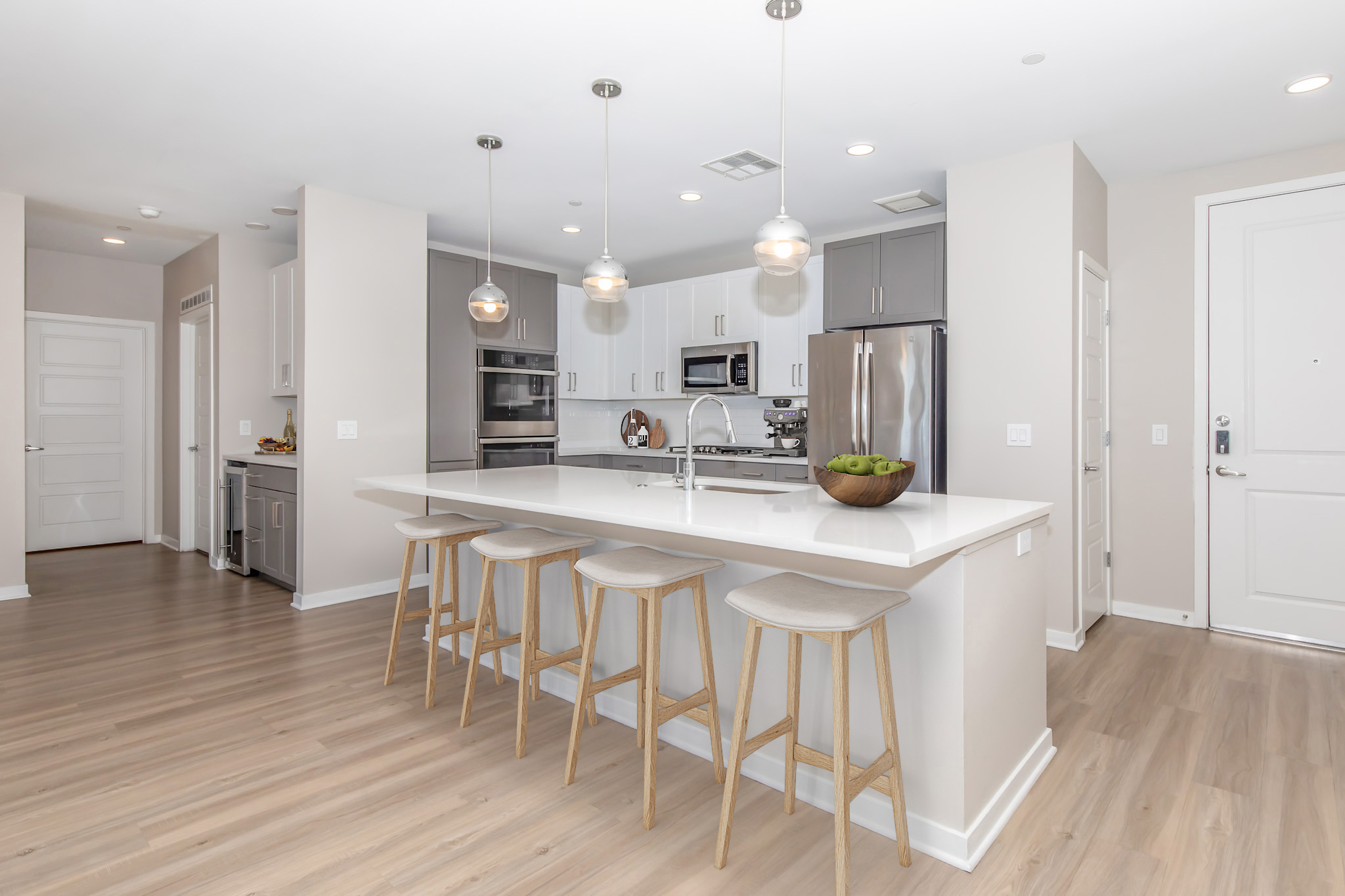 Modern kitchen featuring a large island with stools, stainless steel appliances, gray cabinetry, and pendant lighting. The open layout allows for natural light, enhancing the warm wood flooring and neutral wall colors. The design creates a contemporary and inviting atmosphere.