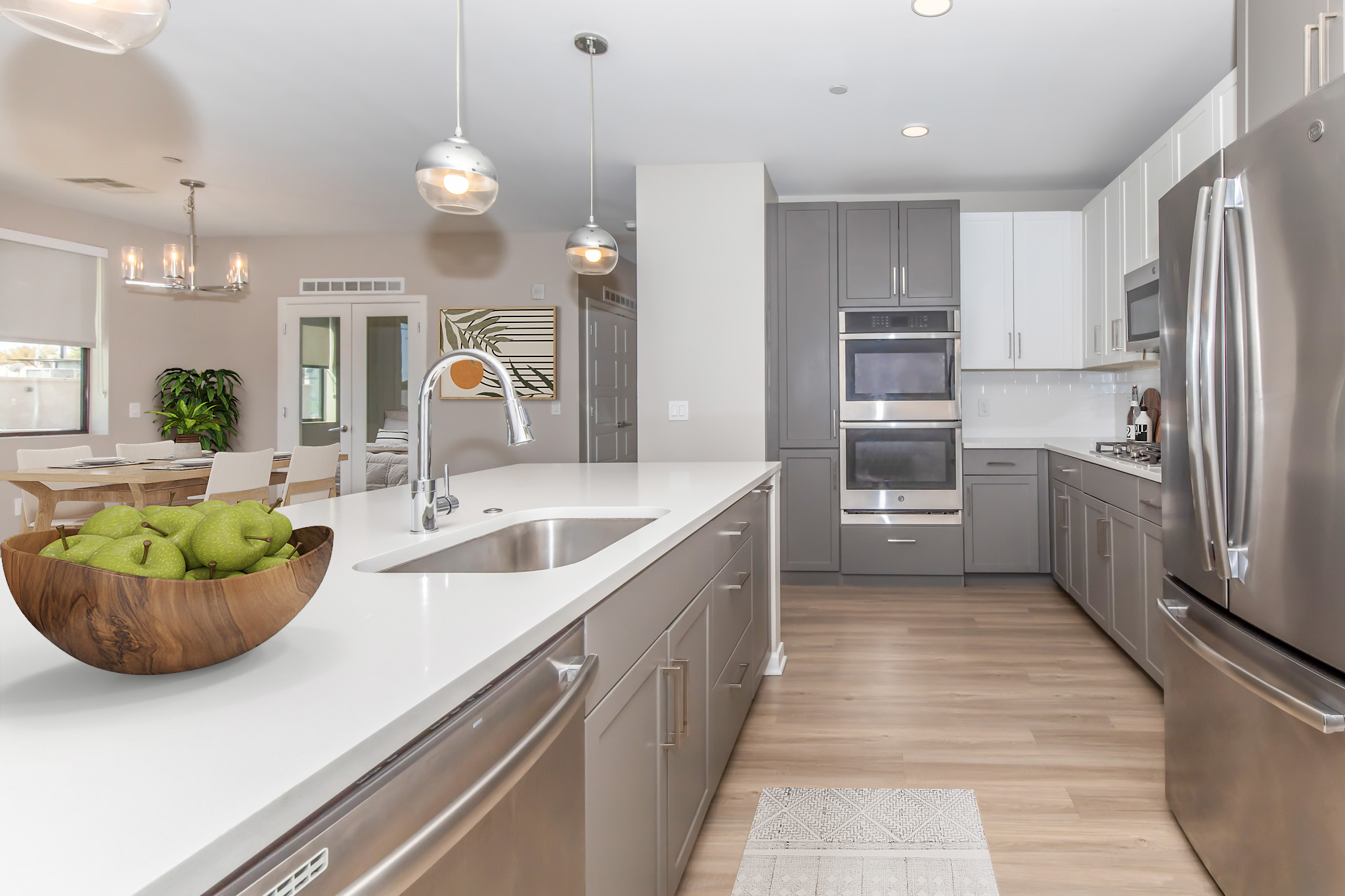 A modern kitchen featuring sleek gray and white cabinetry, stainless steel appliances, and a large island with a white countertop. A bowl of green apples sits on the island, while a dining area is visible in the background, showcasing a light fixture and a wooden dining table.