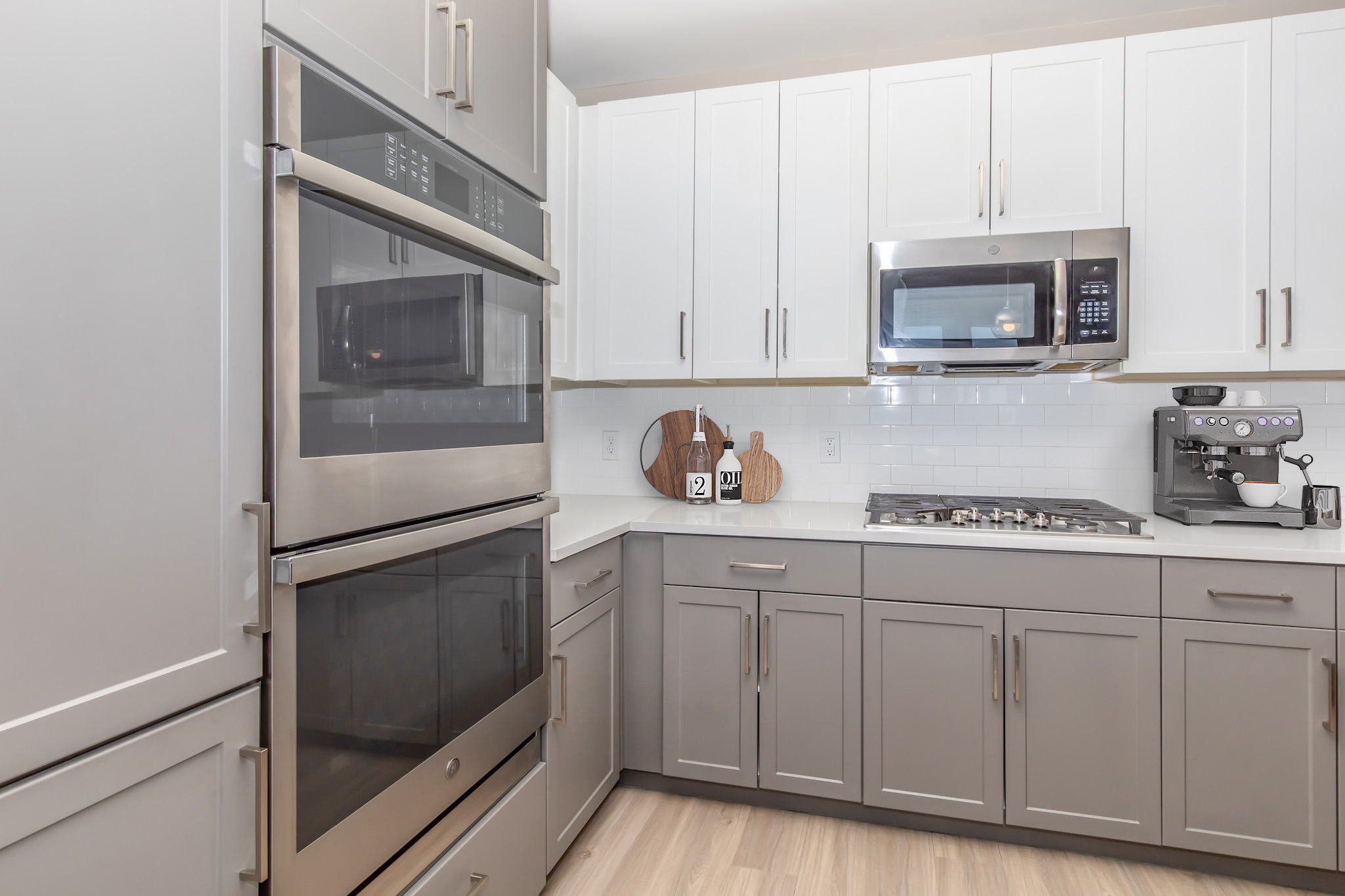 Modern kitchen featuring sleek gray and white cabinetry, stainless steel appliances including a microwave and double ovens, a gas stove, and a coffee maker. The countertops are light-colored, and there are decorative wooden cutting boards on display. The overall design is contemporary and functional.