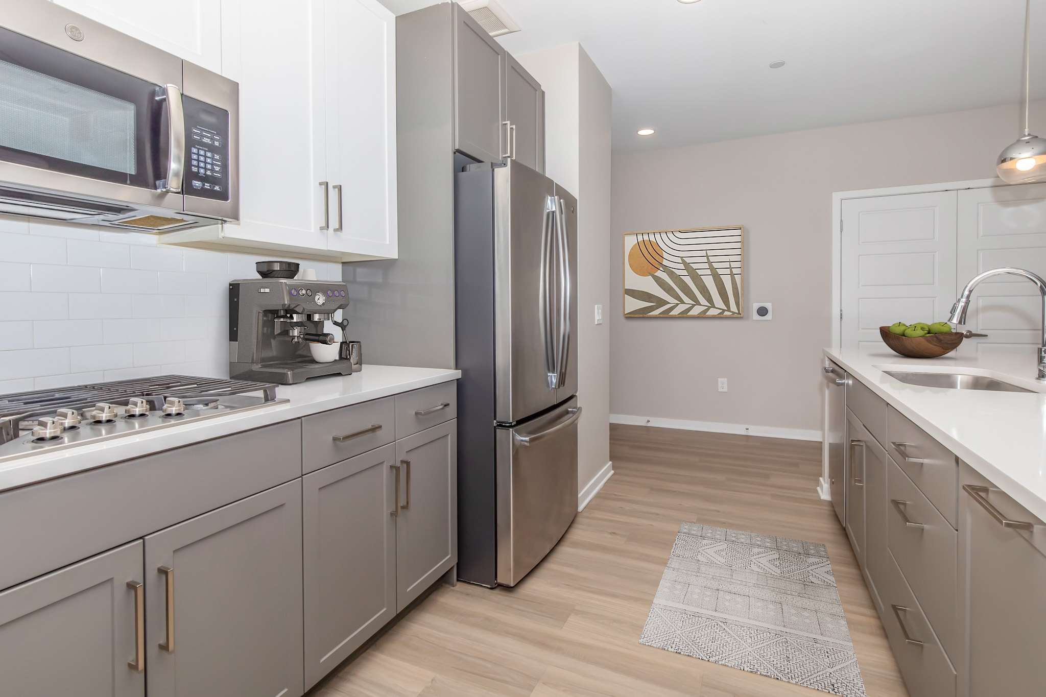 A modern kitchen featuring stainless steel appliances, including a microwave and a sleek dark refrigerator. The cabinetry combines light and dark shades, and there’s a coffee machine on the countertop. Light wood flooring and a rug add a warm touch, with a decorative wall art piece visible in the background.