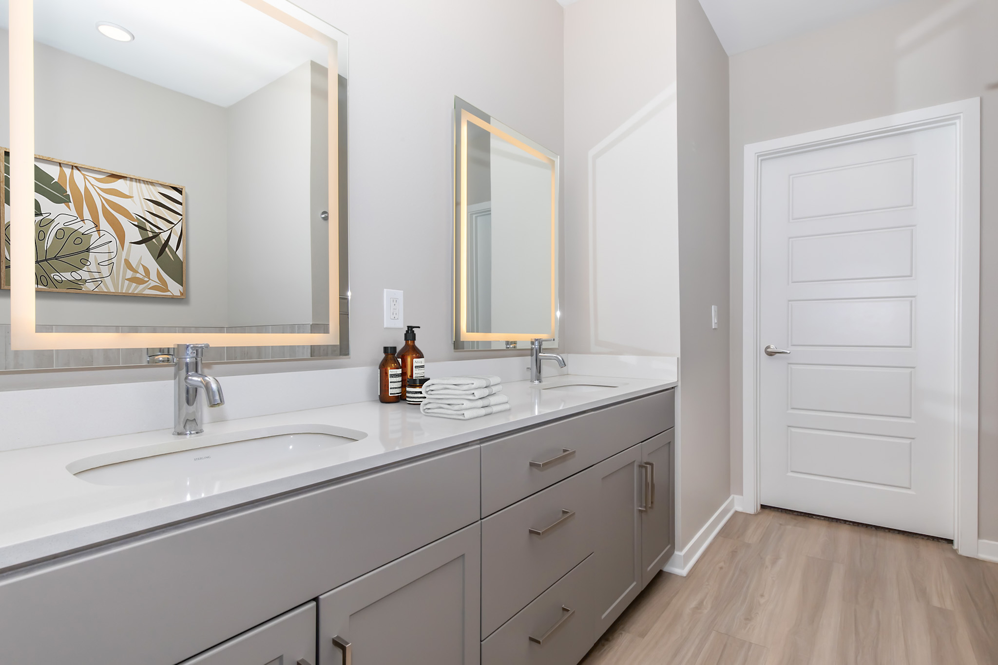 Modern bathroom featuring a sleek double vanity with two sinks, a large mirror illuminated by backlighting, neatly arranged toiletries, and a white door visible in the background. The walls are painted in a light color, and a decorative piece is hanging on the wall.