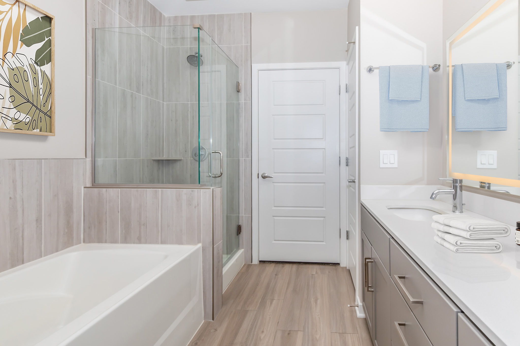 Modern bathroom featuring a glass shower enclosure, a white bathtub, and a double sink vanity with light-colored cabinets. Towels hang on the wall near the mirror, and there is a piece of wall art depicting green leaves. The flooring is light wood, creating a bright and inviting atmosphere.