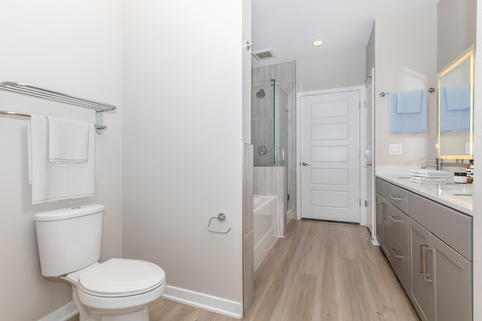 A modern bathroom featuring a toilet on the left, a glass shower enclosure on the right, and a double vanity with a large mirror. The walls are painted in a neutral tone, and the floor is wood-look laminate. Two blue towels hang on a rack, and the space appears clean and well-lit.