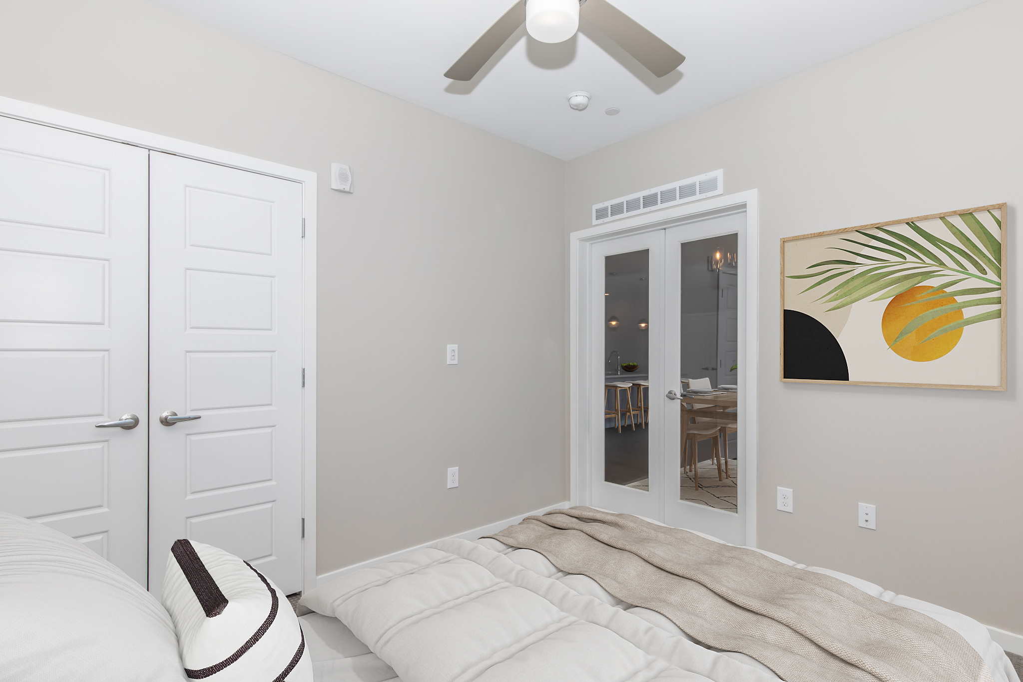 A stylish bedroom with neutral-colored walls, featuring a soft bed with linens, two closed doors, and a large window leading to another room. A contemporary artwork hangs on the wall, depicting abstract shapes in earthy tones, adding a touch of elegance to the space.