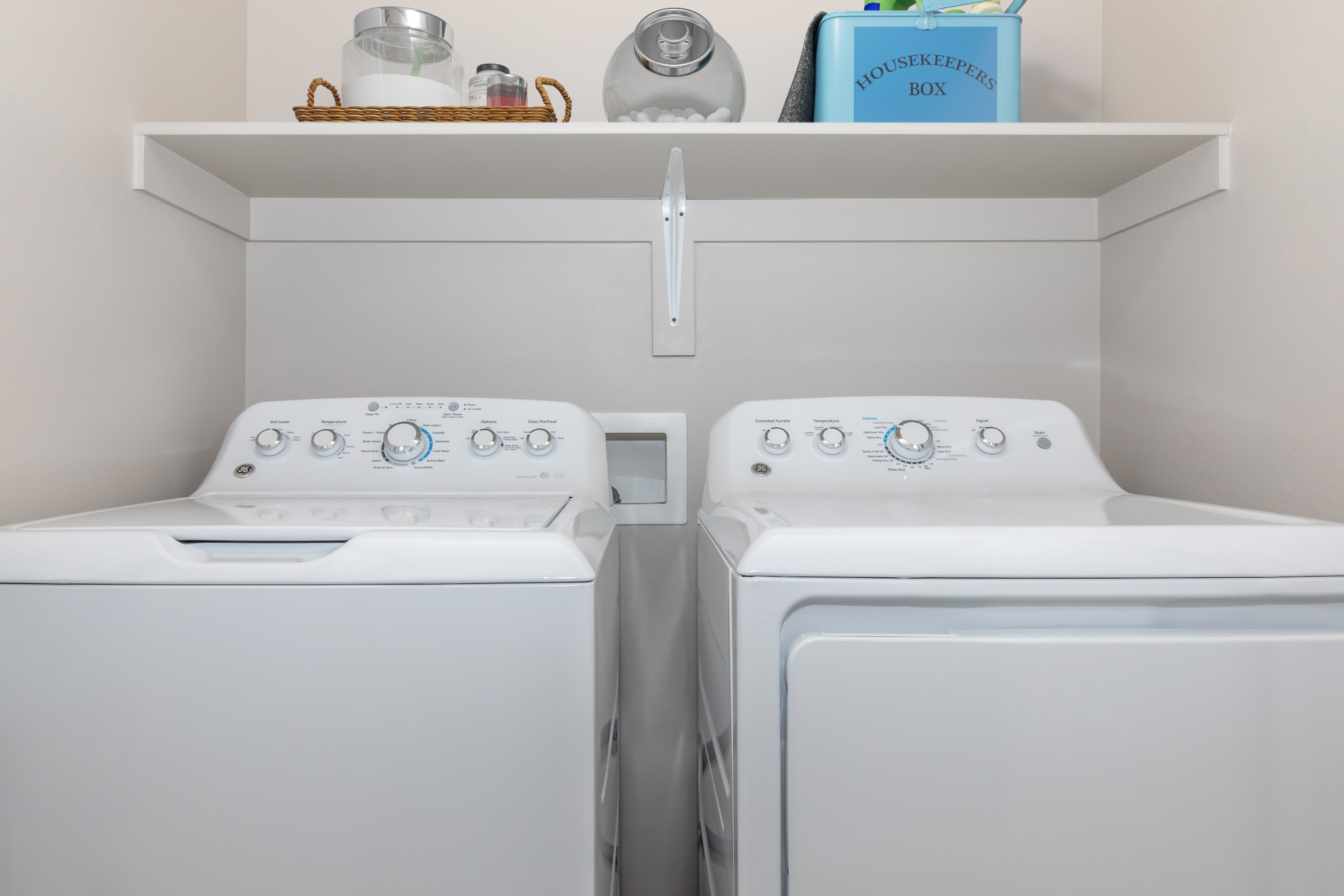 A laundry room featuring a white washing machine and a dryer side by side. Above them is a shelf holding a basket and containers, with a blue box labeled "HOUSEKEEPERS BOX." The walls are light-colored, creating a clean and organized environment.