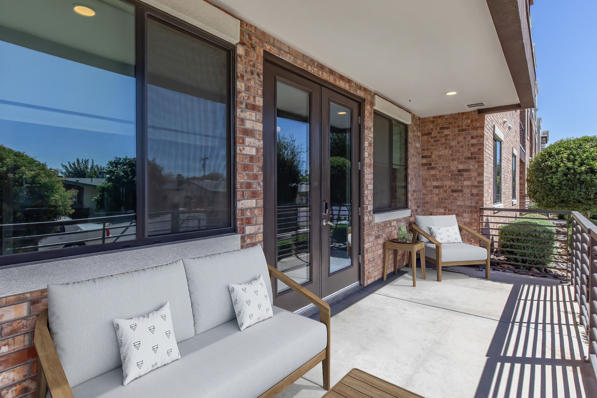 A cozy outdoor patio area features two gray sofas with decorative pillows and a wooden side table. The space is surrounded by brick walls and large windows that let in natural light, while green shrubs provide a touch of nature. The setting is inviting for relaxation and enjoying the outdoors.