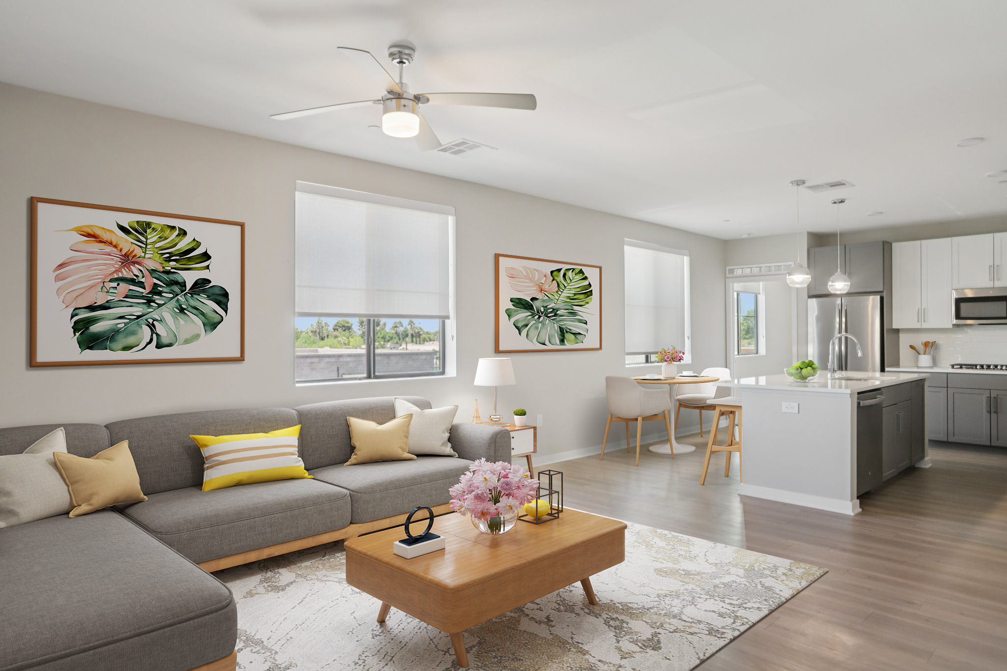 A modern living room and dining area featuring a gray sectional sofa with colorful throw pillows, a wooden coffee table, and a decorative rug. Two framed leaf artwork pieces adorn the walls, and natural light streams in through large windows. A stylish dining table and chairs are visible in the background.