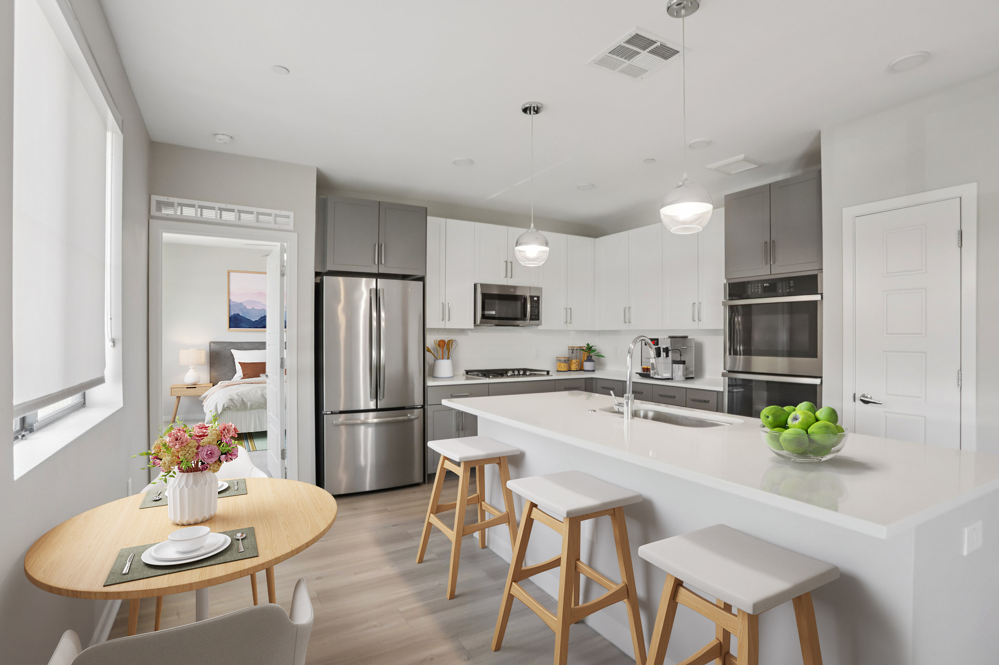 A modern kitchen featuring stainless steel appliances, white and gray cabinetry, and a large island with bar stools. A small dining table is set with plates and a flower vase, while green apples sit in a bowl on the island. A view of a bedroom is visible through an open door.