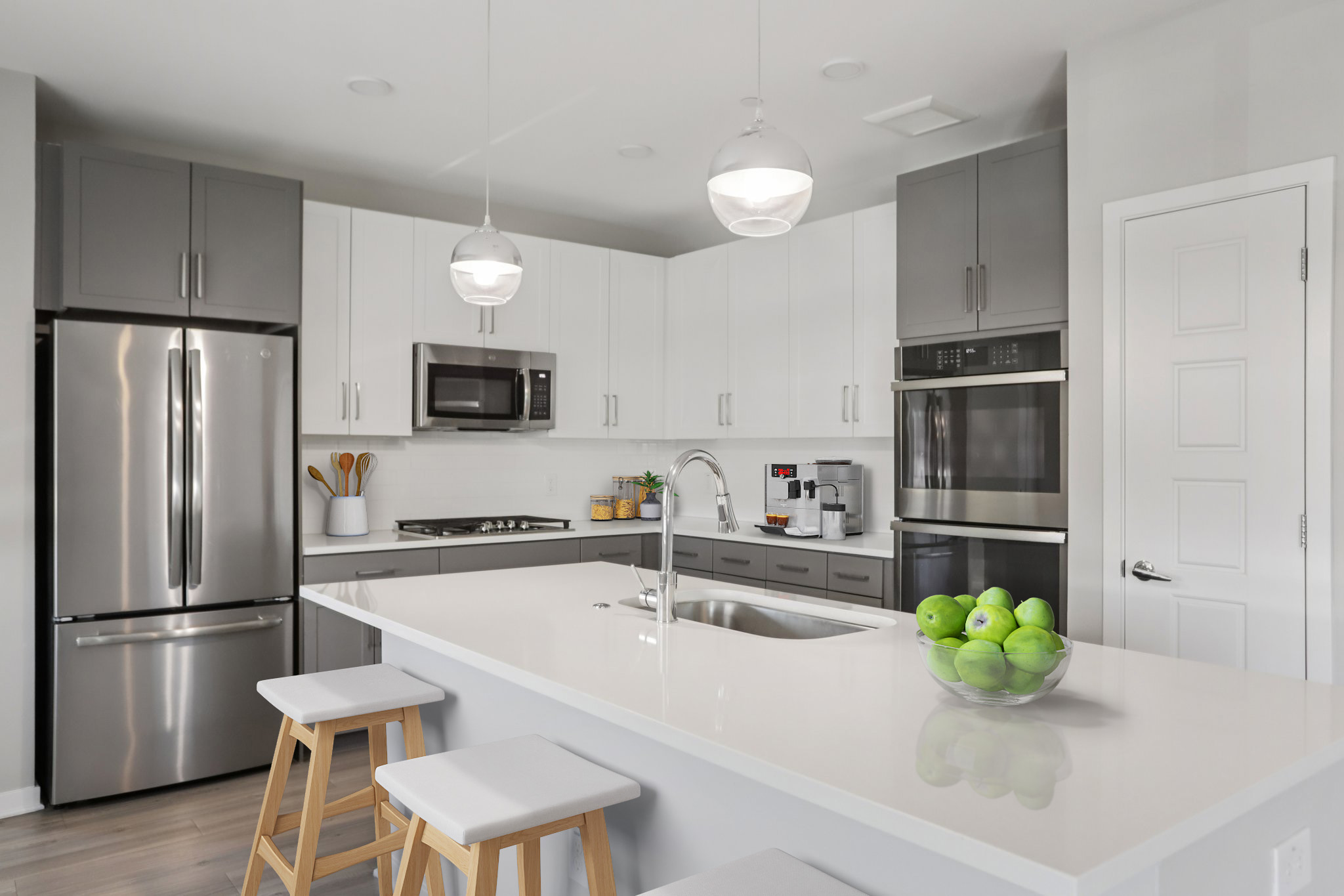 Modern kitchen featuring a spacious island with three stools, stainless steel appliances, a microwave, and dual ovens. The cabinetry is a mix of dark and light colors, and a bowl of green apples is displayed on the countertop. Pendant lights hang above the island, enhancing the contemporary design.
