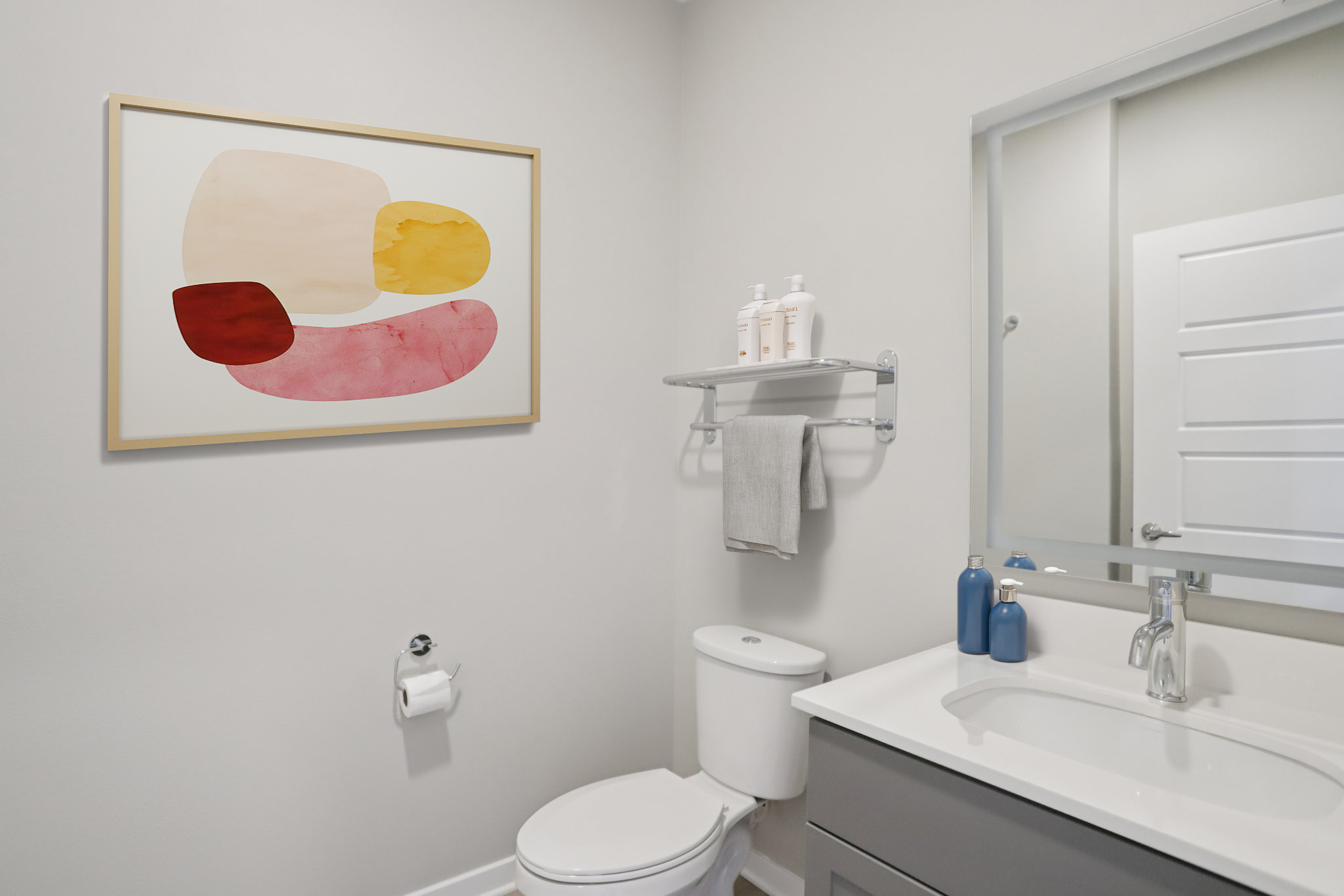 A modern bathroom featuring a toilet, a sink with a mirror above it, and a wall-mounted shelf holding toiletries. There is a colorful abstract art piece framed on the wall. The walls are painted in a light shade, and the decor is minimalistic and clean.