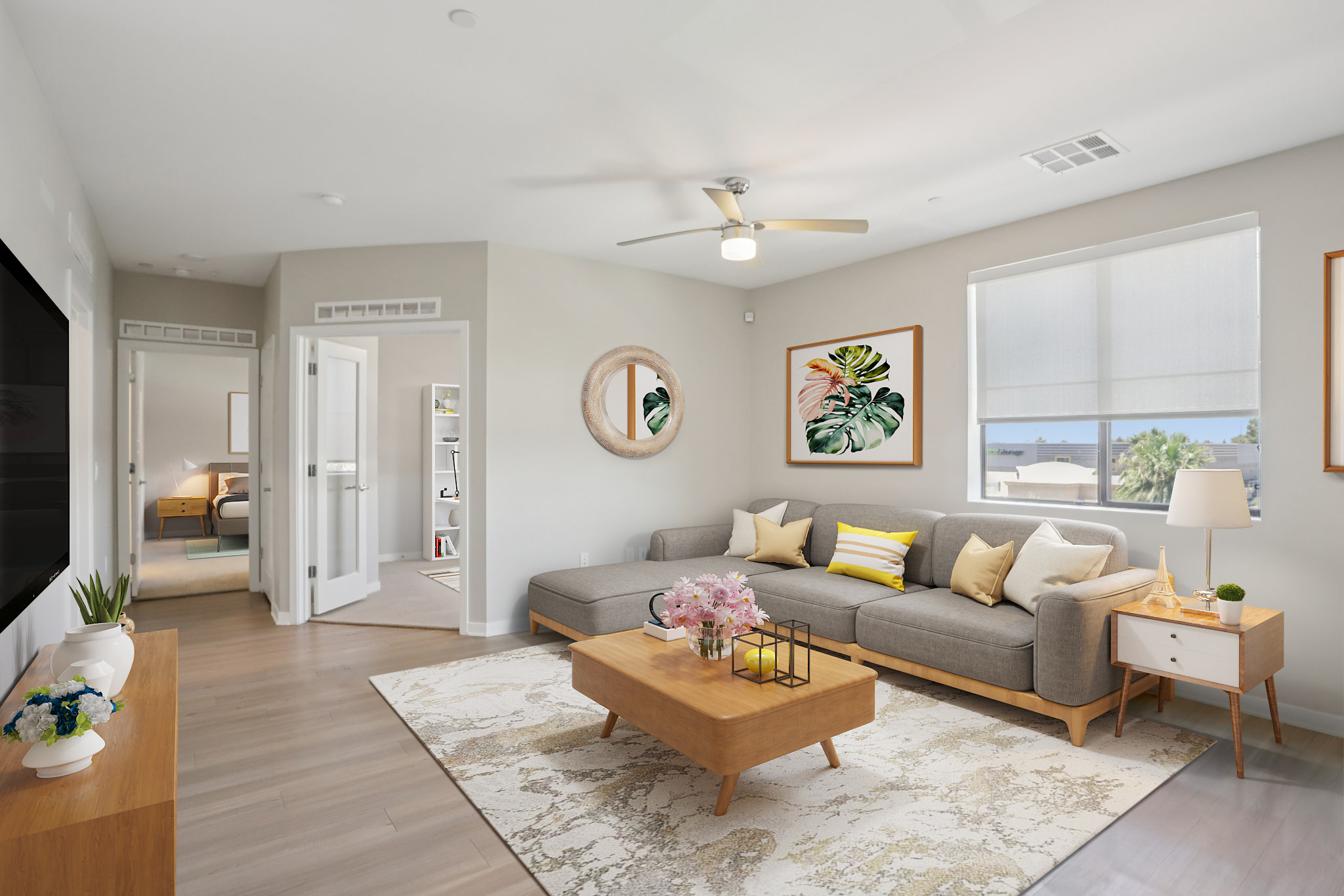 A modern living room featuring a gray sectional sofa adorned with yellow and white cushions. A wooden coffee table sits in the center, topped with flowers. The walls display framed botanical art. A large window lets in natural light, and a ceiling fan hangs above. The atmosphere is bright and inviting.
