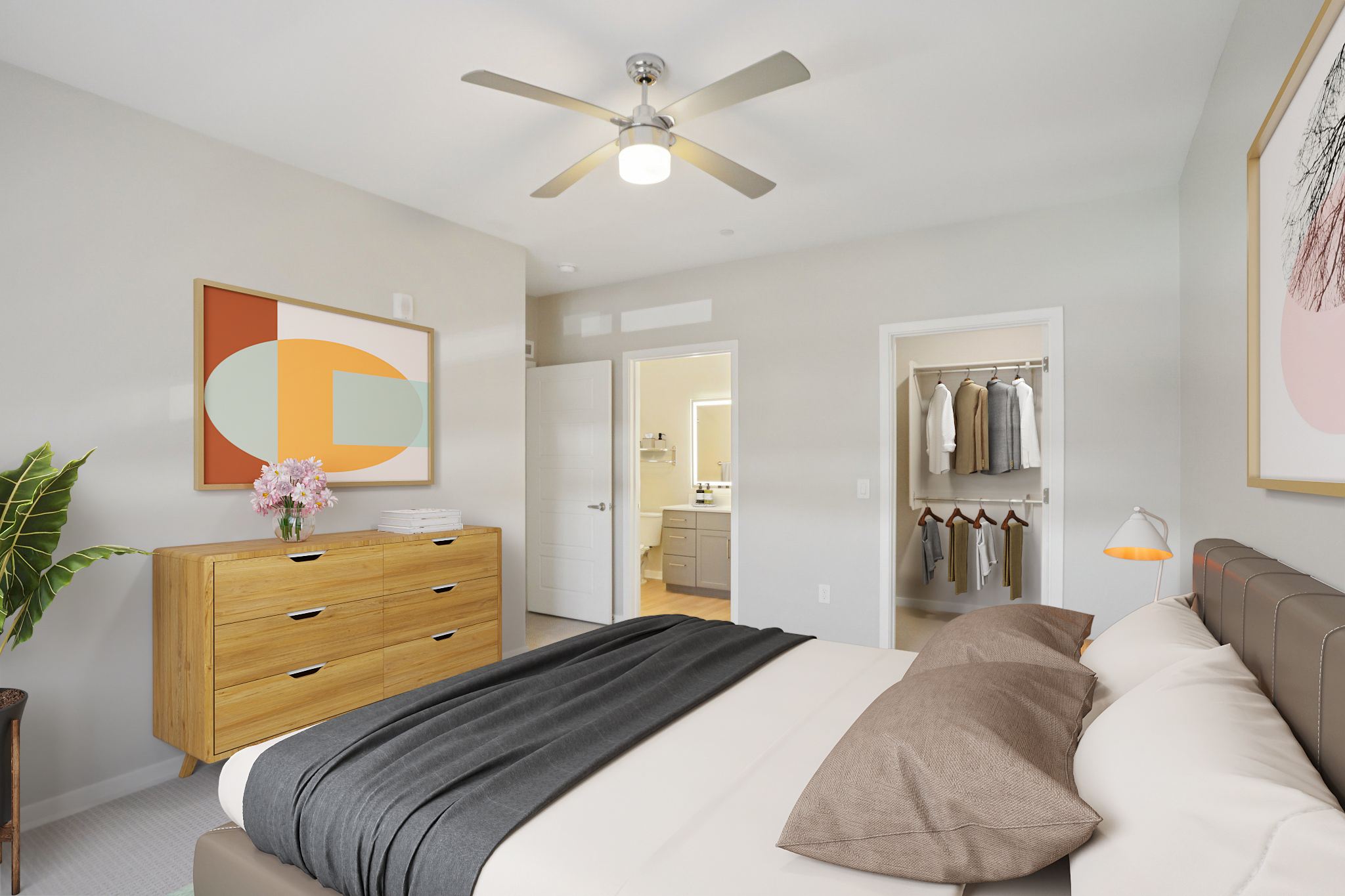 A modern bedroom featuring a comfortable bed adorned with brown pillows and a dark throw blanket. There is a wooden dresser and a decorative plant, along with an abstract painting on the wall. A door leads to a bathroom, while a closet shows neatly hung clothes. The room has a ceiling fan and natural light.