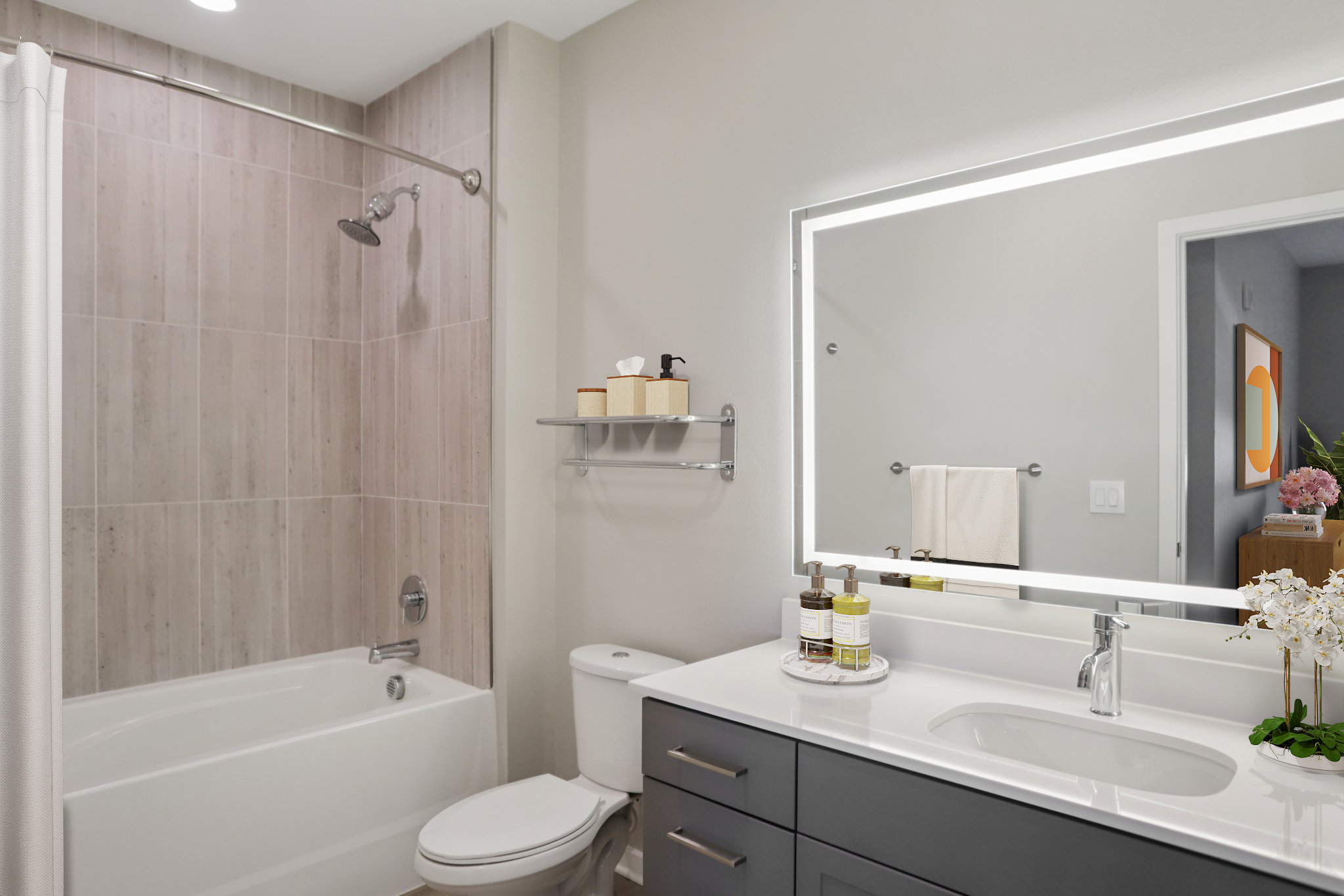 Modern bathroom featuring a white bathtub with a shower, a stylish sink with a dark cabinet, and a large mirror with backlighting. The walls are light-colored, and there are decorative items on the countertop, including bottles and an orchid plant, creating a clean and inviting atmosphere.