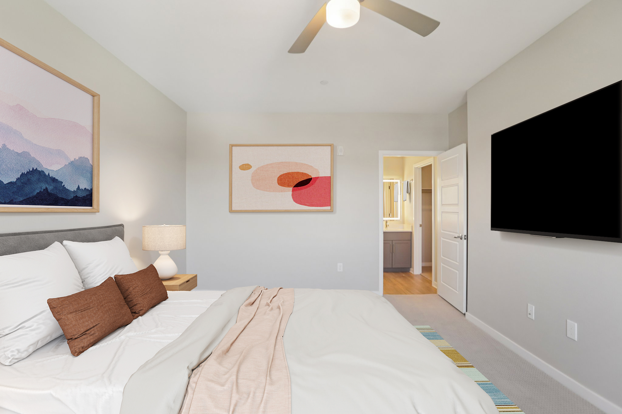 A modern bedroom featuring a gray bed with white linens and brown accent pillows. The room has two framed artworks on the walls, a bedside lamp, and a large flat-screen TV mounted on the wall. A doorway leads to a bathroom, and the floor is covered with a colorful rug. Natural light brightens the space.