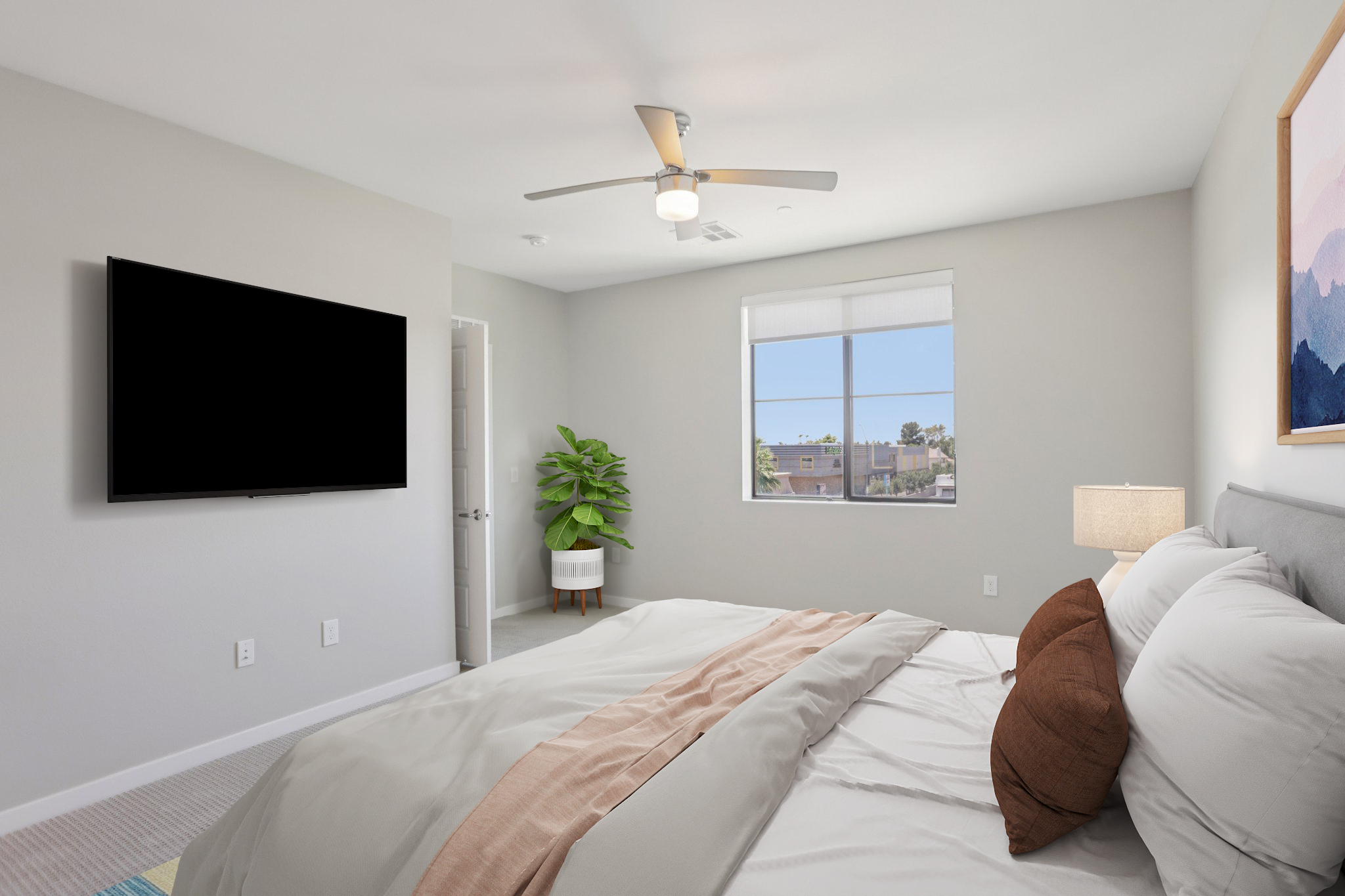 A modern bedroom featuring a neatly made bed with light bedding and decorative pillows, a ceiling fan, a large wall-mounted TV, a bedside lamp, and a potted plant. The room has neutral walls and a window with a view.