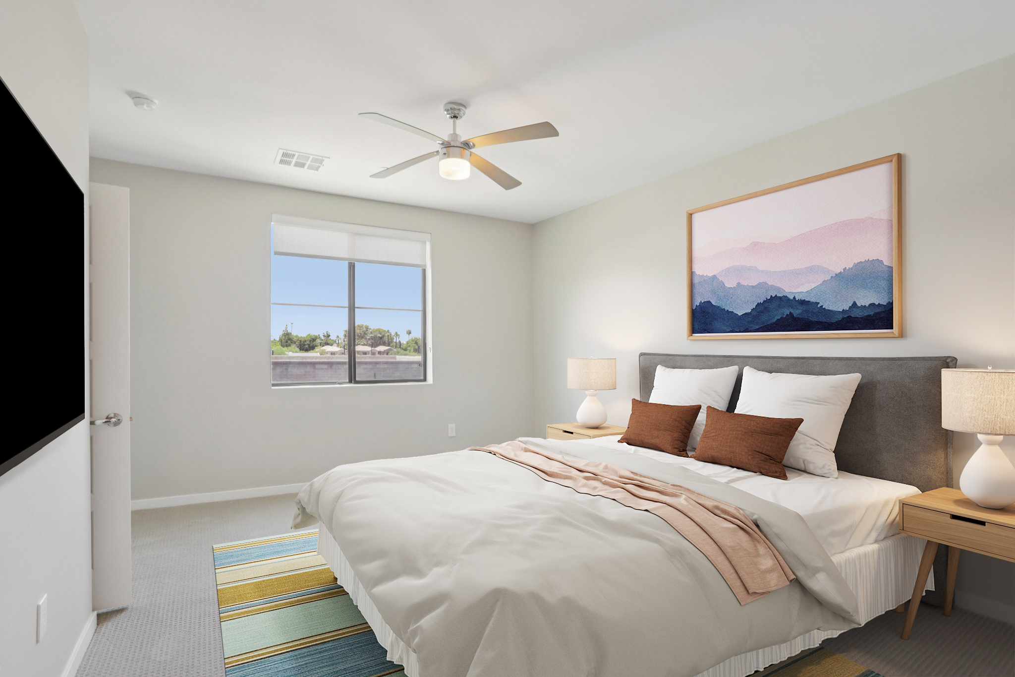 A cozy bedroom featuring a comfortable bed with white and beige bedding, two brown accent pillows, bedside tables with lamps, a large window with a view, and a wall-mounted TV. A landscape artwork hangs above the bed, and the room is decorated with light gray walls and a colorful striped rug on the floor.