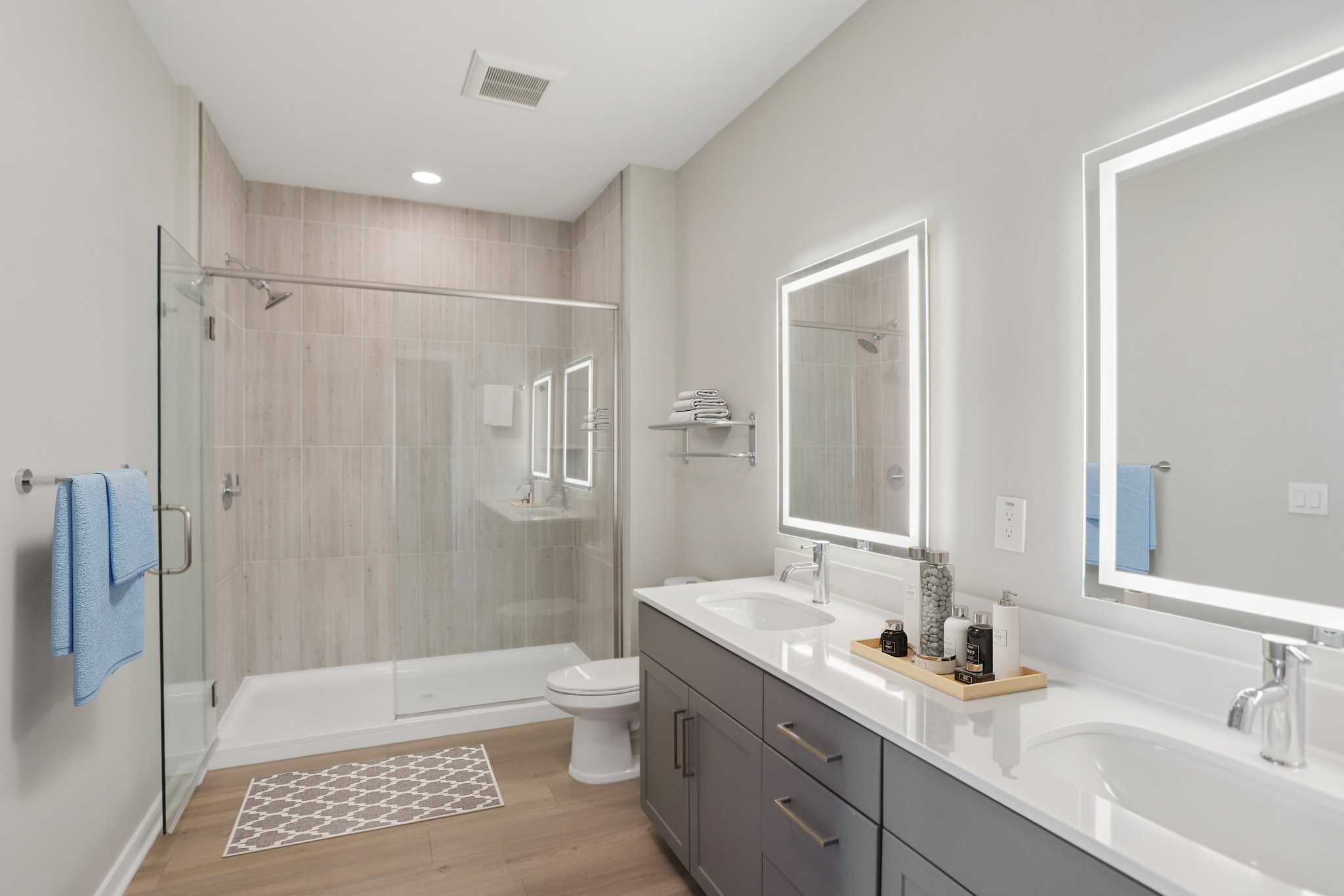 Modern bathroom featuring a glass shower enclosure, dual sink vanity with under-mount sinks, sleek gray cabinetry, and illuminated mirrors. The space is adorned with light wood-style flooring, a blue towel hanging on the wall, and a decorative rug on the floor.