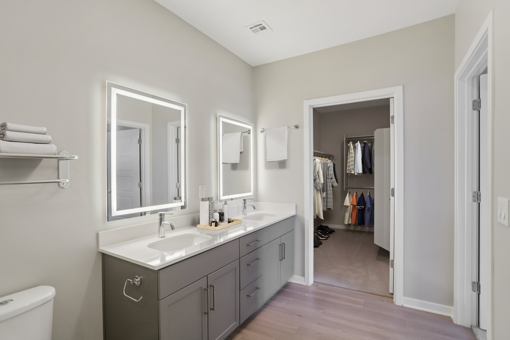 Modern bathroom featuring dual sinks with backlit mirrors, a sleek countertop, and a towel rack. The design includes light gray walls, and a doorway leading to a closet with organized clothing. The flooring is a light wood tone, creating a contemporary and spacious atmosphere.