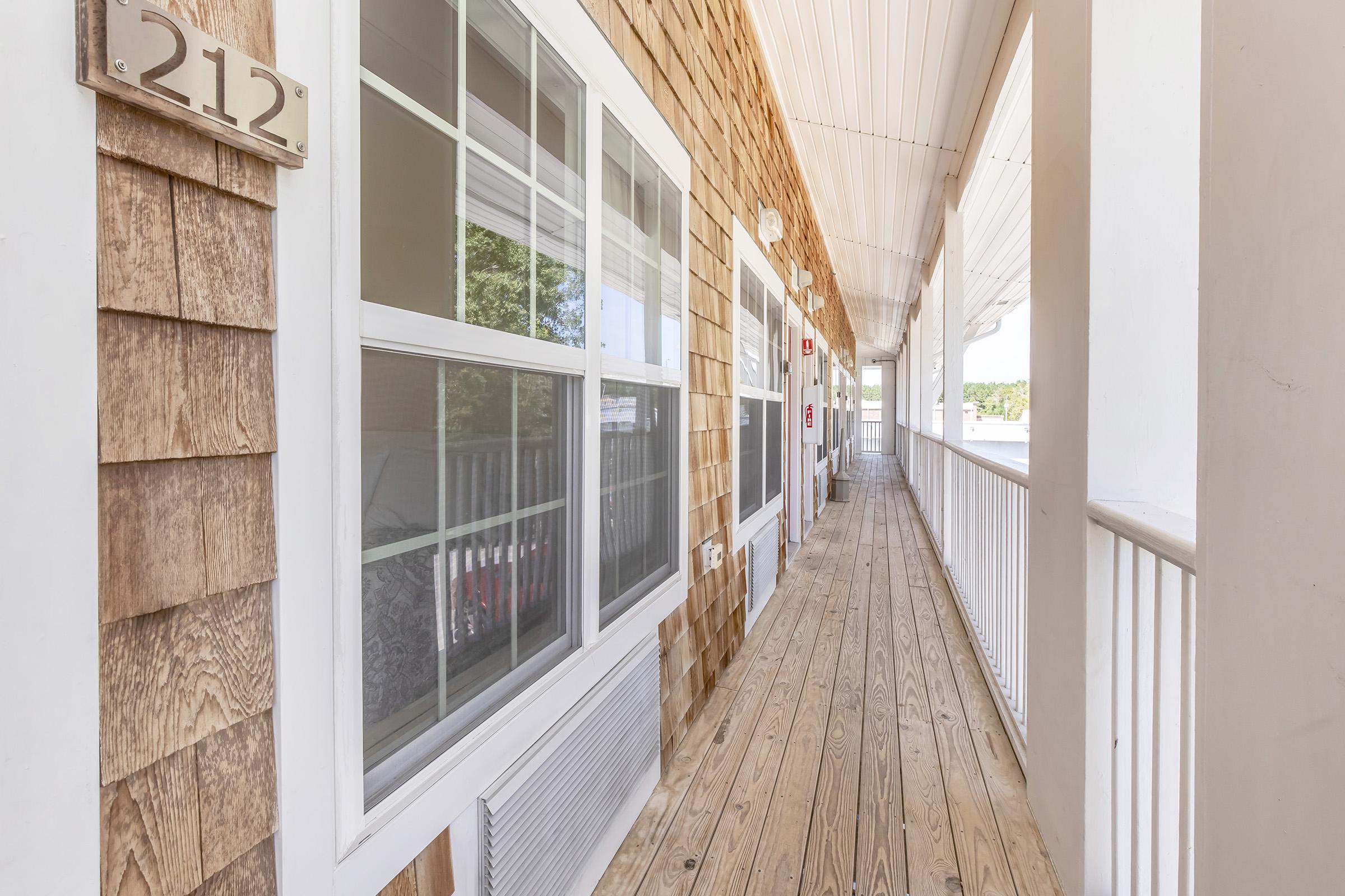 View of a hotel corridor featuring wooden siding and a long, wooden floor. The hallway includes several doors, one labeled "212." Bright natural light shines through the windows, creating an inviting atmosphere.