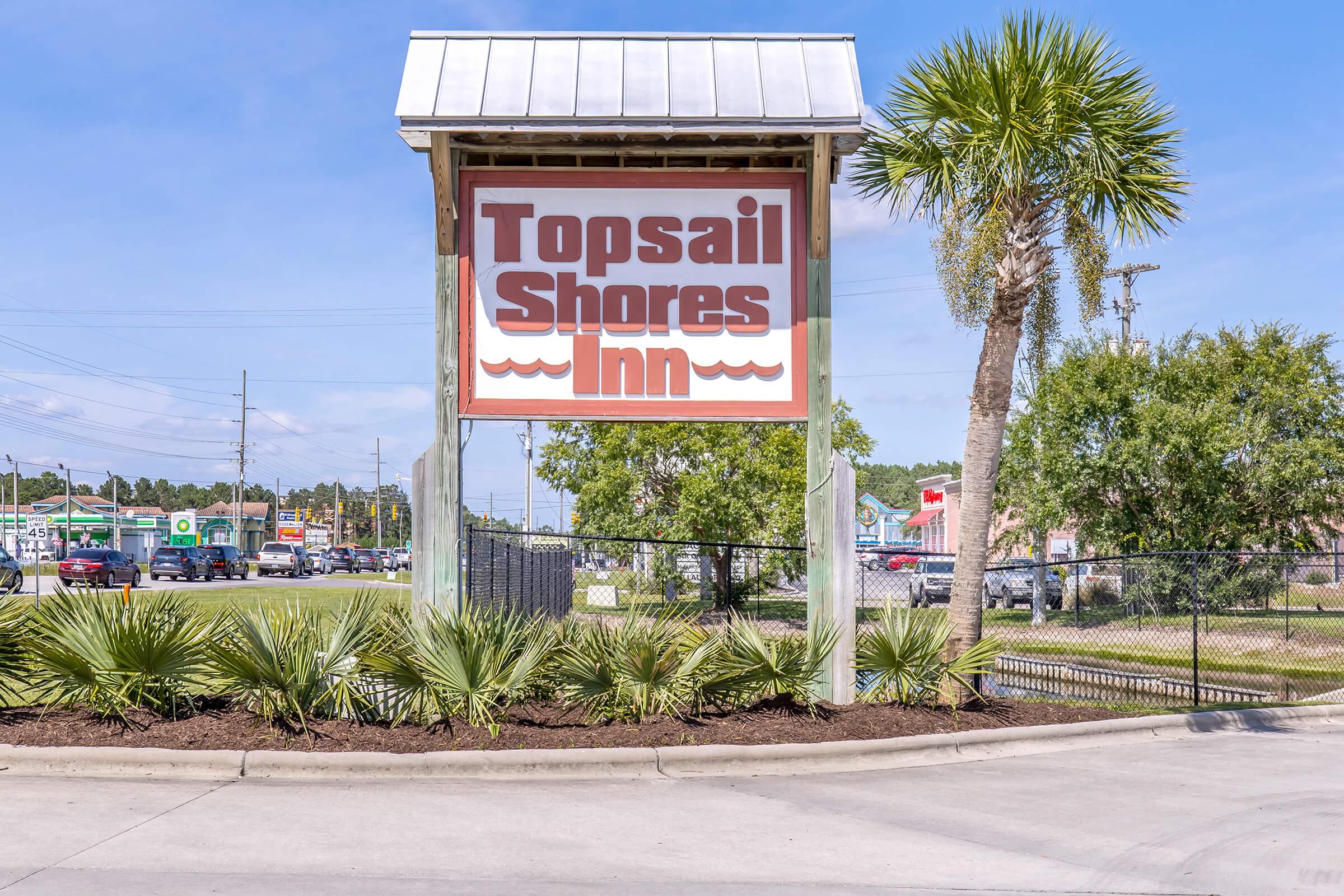 Sign for Topsail Shores Inn, featuring a large, bold font with waves depicted beneath the text. Surrounding the sign are palm trees and shrubs, with a clear sky in the background. The image shows a parking area and nearby businesses in the vicinity.